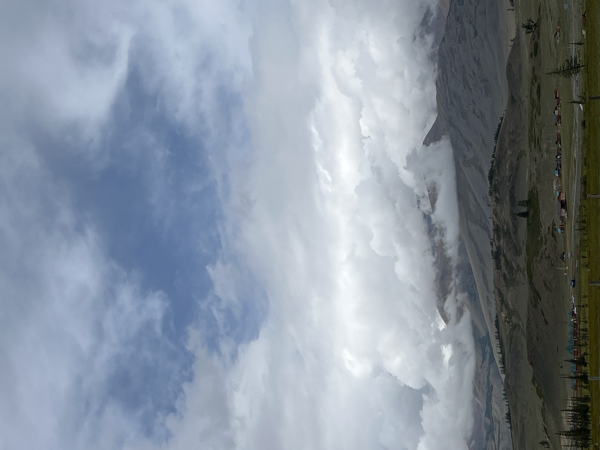 A wide, vertical shot captures a mountainous landscape under a dramatic sky. The upper portion of the image is dominated by a layered sky, with lighter, wispy clouds transitioning into heavier, more substantial formations that obscure much of the blue. These clouds are dense and appear to be gathering around the peaks of the mountains in the midground.
The mountains themselves are rugged and appear to be covered in sparse vegetation. Slopes are visible, with some areas appearing lighter and others darker, suggesting varied terrain and possibly shadows. The lower portions of the mountains descend into rolling hills.
In the foreground, a line of small, red-roofed structures with green trim are visible, arranged in a semi-circle. These appear to be dwellings or cabins. Several trees, some tall and slender, are interspersed between and around these structures. A grassy plain stretches out in front of the structures, with a road or path running through it. The overall impression is of a remote, possibly rural or nomadic settlement situated in a vast, natural environment. The location is identified as Kyzyl-Tash, Russia. There are no people or animals clearly visible in the image.