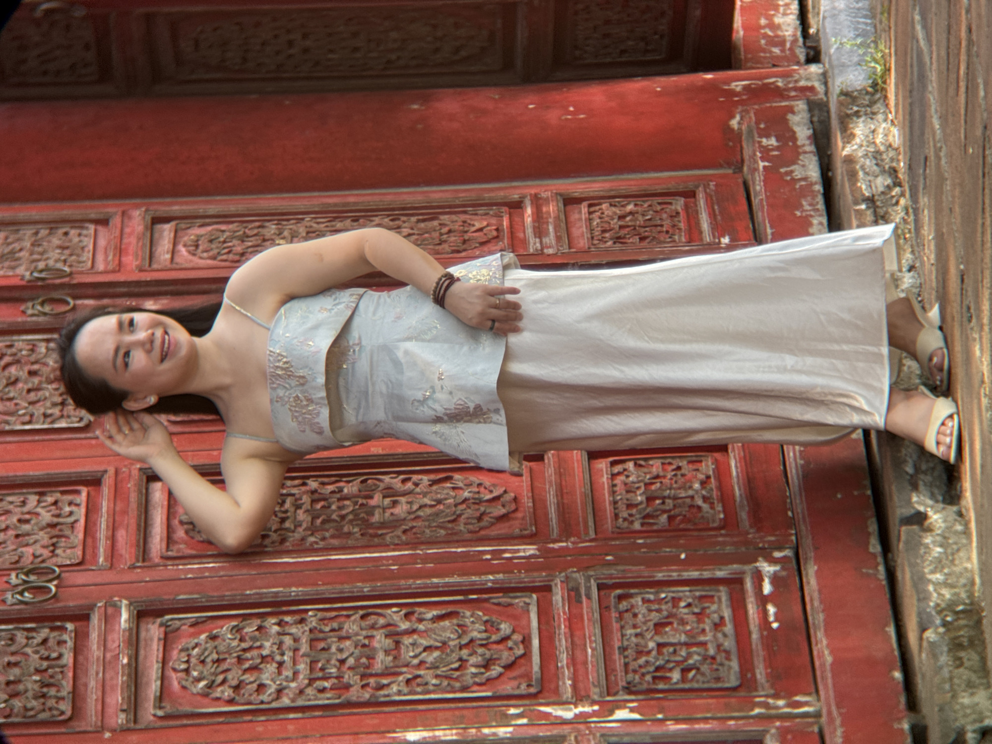 A Vietnamese woman stands in front of a red, ornately carved wooden door, likely at the Imperial City of Hue in Phú Xuân Ward, Vietnam. She wears a light-colored, tiered dress with a fitted peplum and a flowing skirt, adorned with subtle embroidery. Her hair is pulled back, and she has a beaded bracelet on her wrist and a ring on her finger. She is smiling and has one hand raised near her ear, possibly adjusting her hair or shielding her eyes. The door has decorative carvings and metal rings. The ground is paved with stone and concrete, with some greenery visible.