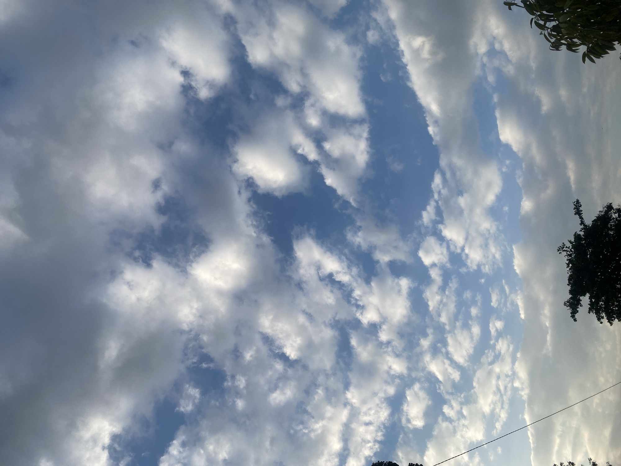 The image is an outdoor photograph taken in Banjarbaru, Indonesia, showcasing a sky filled with scattered clouds against a bright blue backdrop. The clouds vary in size and density, with some appearing wispy and others more substantial and textured. The sunlight filters through the clouds, creating areas of light and shadow. In the lower portion of the frame, the dark silhouettes of trees and a thin, dark wire are visible, suggesting the presence of vegetation and possibly a power line. The overall scene depicts a natural environment with a dynamic sky.