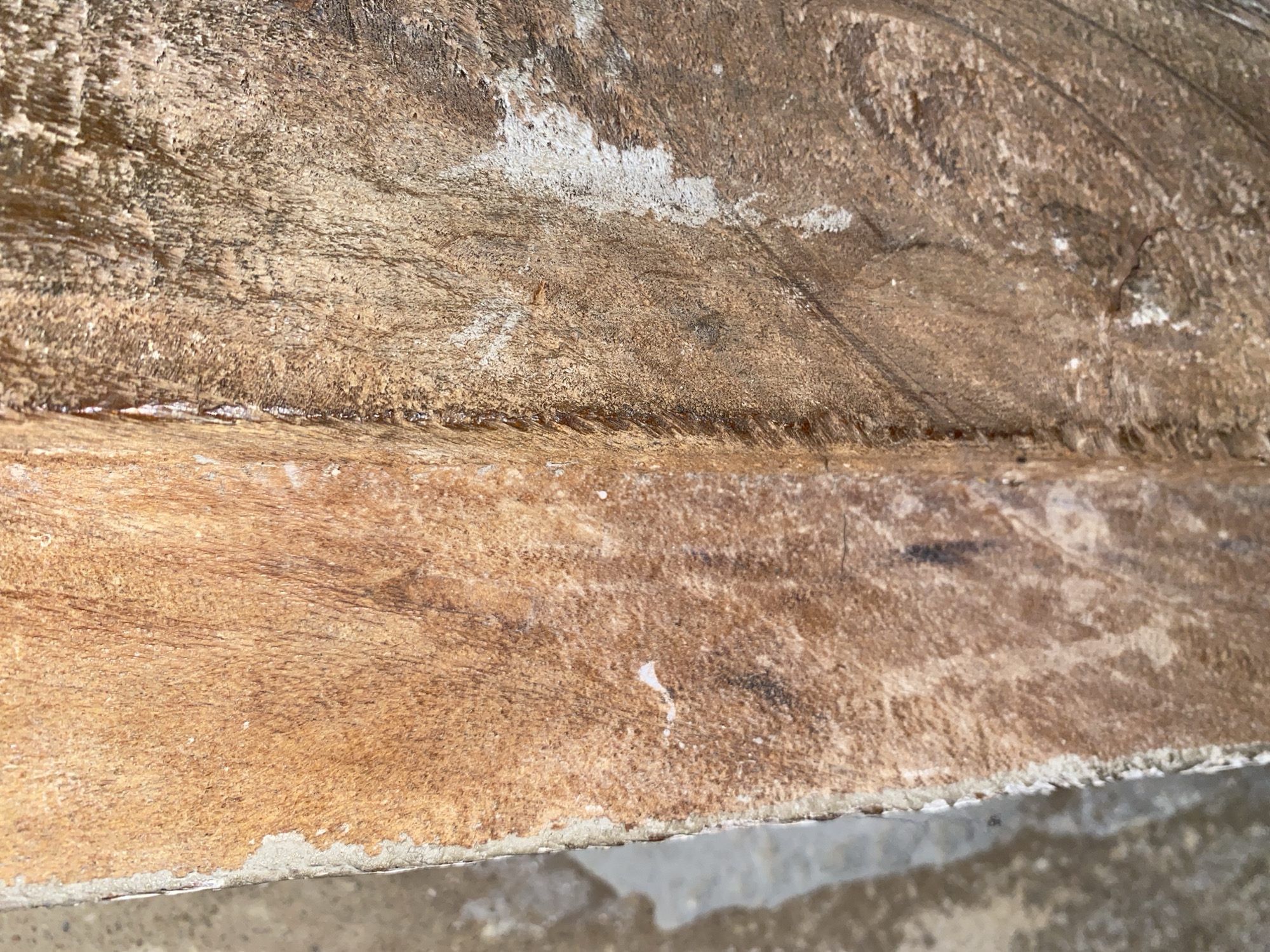 The image displays a close-up view of weathered wooden planks, likely part of a structure or object in Jauro Yinu, Nigeria. The wood exhibits a rough, textured surface with visible grain patterns and variations in color, ranging from light brown to darker, aged hues. Some areas show signs of water damage or staining, with lighter, almost white patches that could be mineral deposits or mildew. The edge of the wood on the left side appears to have some gray, gritty material adhering to it, possibly concrete or a similar building material. The lighting suggests an outdoor setting, with shadows and highlights indicating a light source. The image does not contain any people or discernible actions, focusing solely on the material and its condition.
