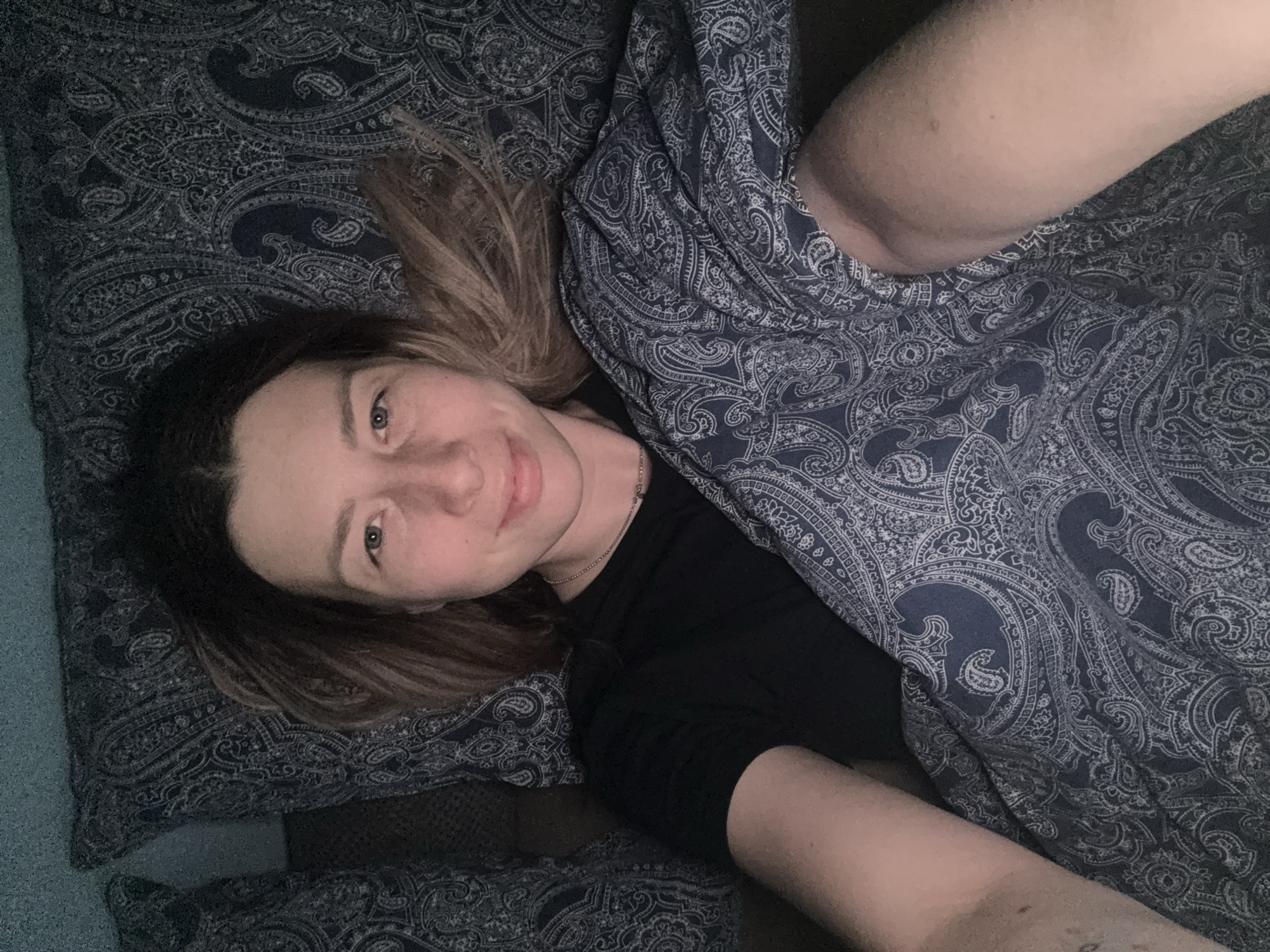 A woman with light brown hair is lying in bed, smiling and looking at the camera. She is wearing a black t-shirt and a silver necklace. She is partially covered by a paisley patterned duvet. The bed has a matching paisley patterned pillow. The location is not specified and cannot be determined from the image.