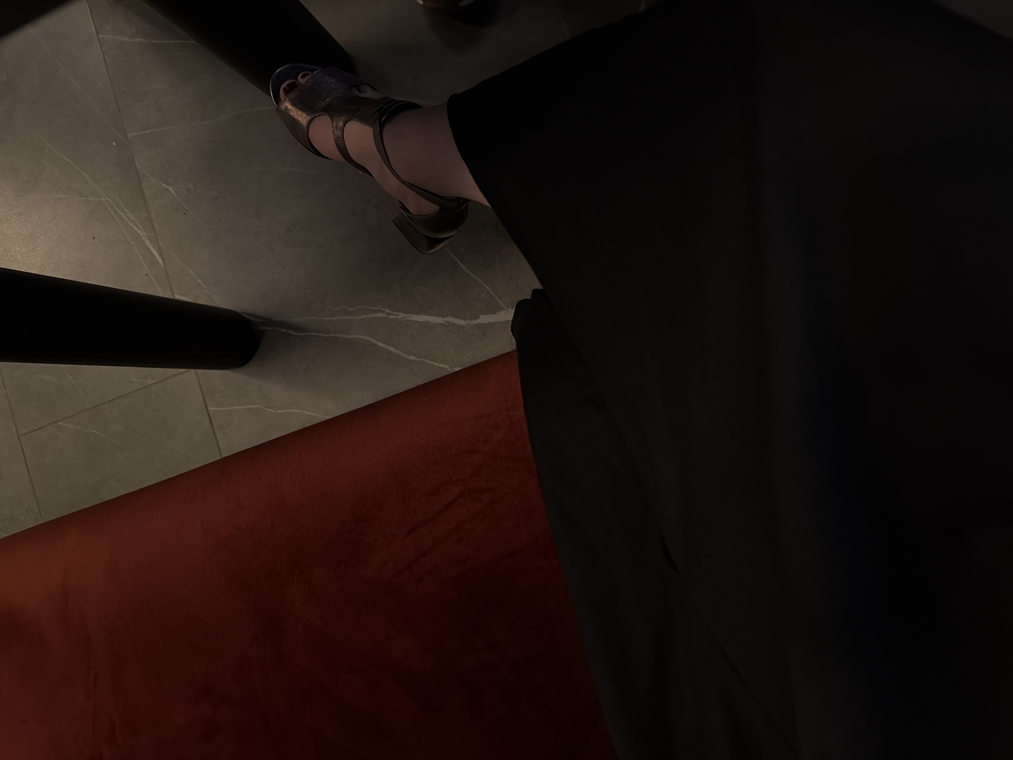 The scene is a dimly lit interior, showing a person seated on an orange velvet surface. The person wears black clothing and metallic gladiator-style heeled sandals. The background features a grey, patterned floor. A black table leg is visible. The location and specific context related to the city of Izhevsk, Russia, are not evident from the image.