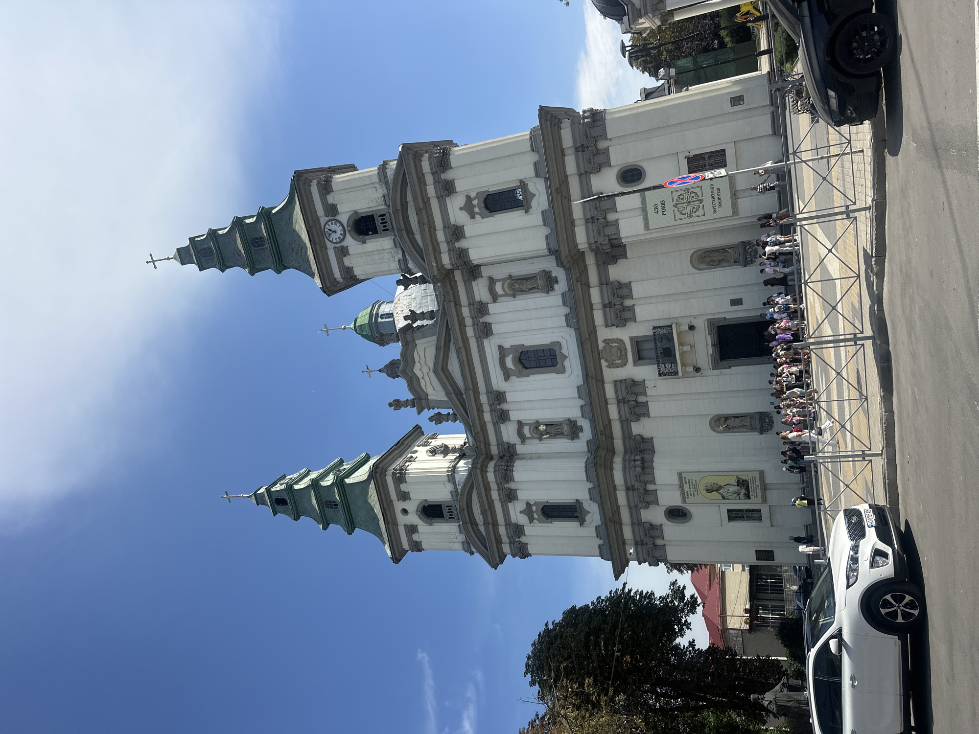 The image shows the Ternopil Cathedral of the Immaculate Conception of the Blessed Virgin Mary in Ternopil, Ukraine. The large, white Baroque-style church features two tall green spires topped with crosses, as well as a clock on one of the towers. The building has ornate white stonework with grey accents, and several statues and architectural details are visible. In front of the church, a group of people are gathered, appearing to be tourists or visitors. A white car is parked on the left side of the frame, and a black car is visible on the right. The sky is blue with some light clouds. A sign with Ukrainian text is mounted on the church wall. The overall scene depicts a sunny day in a European city.
