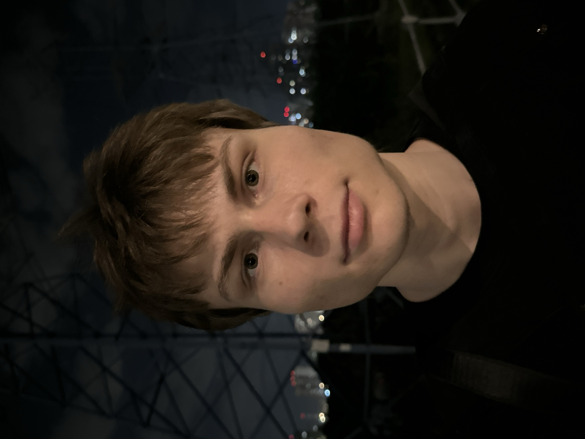 A portrait of a person with light skin, brown hair, and green eyes. The person is wearing a black shirt. The background shows a nighttime urban setting with blurry lights that suggest a city skyline. The location appears to be in Moscow, Russia due to the style of the skyline and architectural elements.