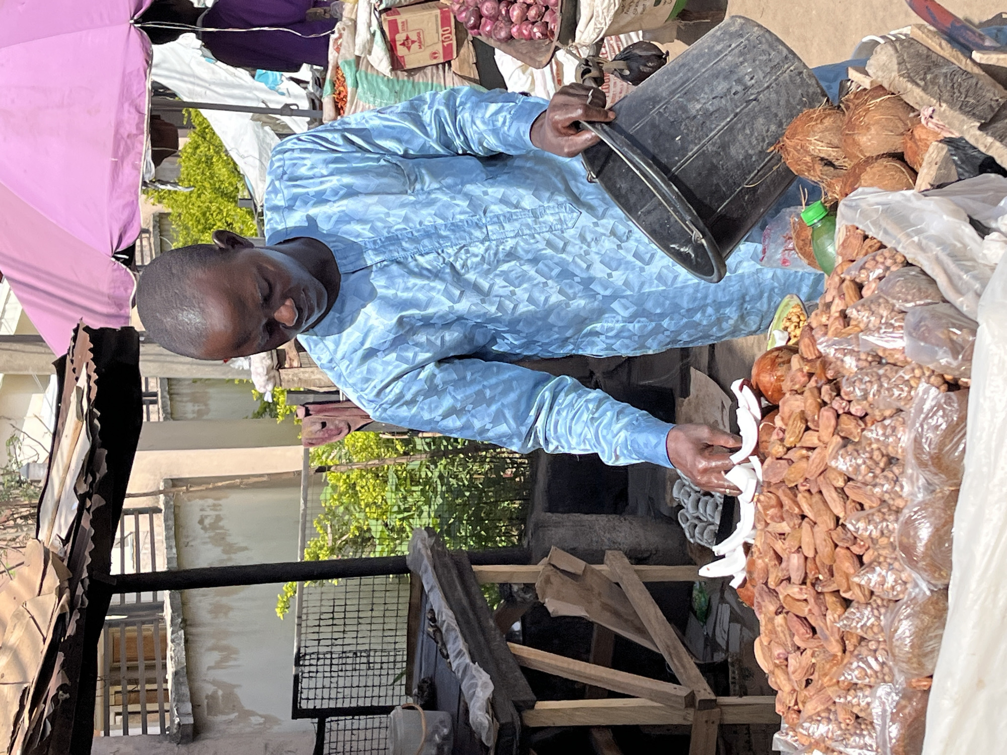 In a marketplace in Jos, Nigeria, a man wearing a light blue, patterned long-sleeved shirt stands behind a stall piled high with goods. He is using a black bucket, possibly to organize or measure items. The stall contains coconuts, bags of nuts, and other produce. Above the stall is a pink umbrella and other items, including onions in bags. The stall is constructed of rough wooden frames. The environment includes buildings, greenery, and a mesh fence, suggesting an urban setting.