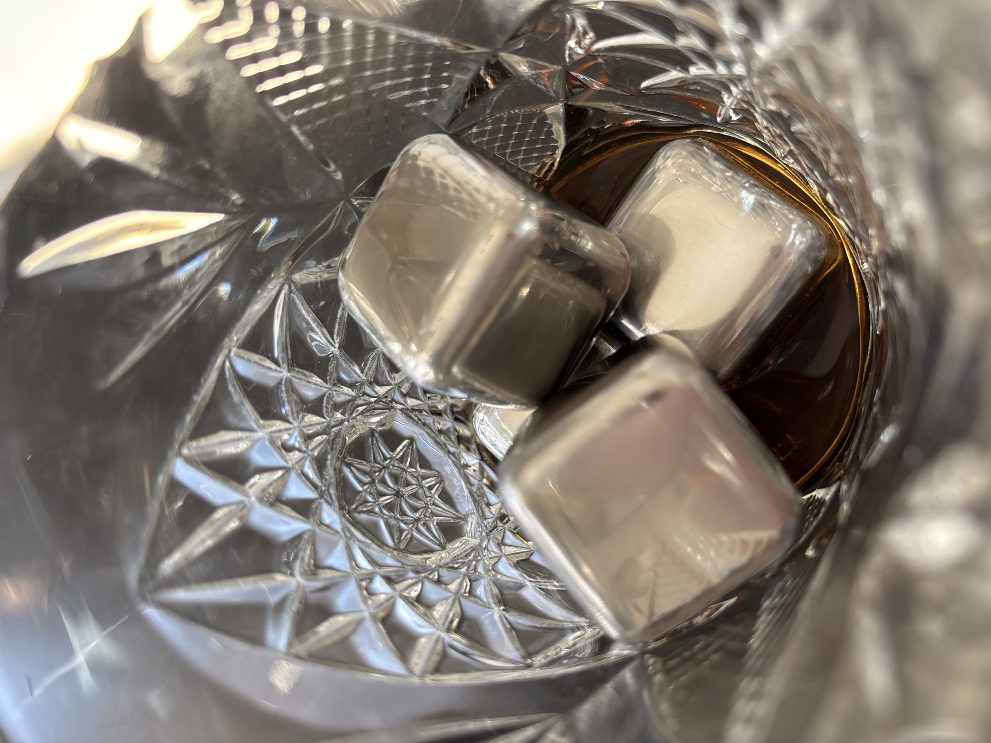 A close-up, overhead shot shows three stainless steel whiskey stones resting in a cut glass tumbler containing amber liquid. The tumbler has a diamond-cut pattern, and light glints off the facets and the stones. The liquid appears to be a spirit such as whiskey or brandy. The image is focused on the contents of the glass, with the rim of the tumbler visible in the upper portion of the frame. There are no people or other objects visible in the scene. The location of Razdolnoye, Russia is not depicted or suggested by the image.