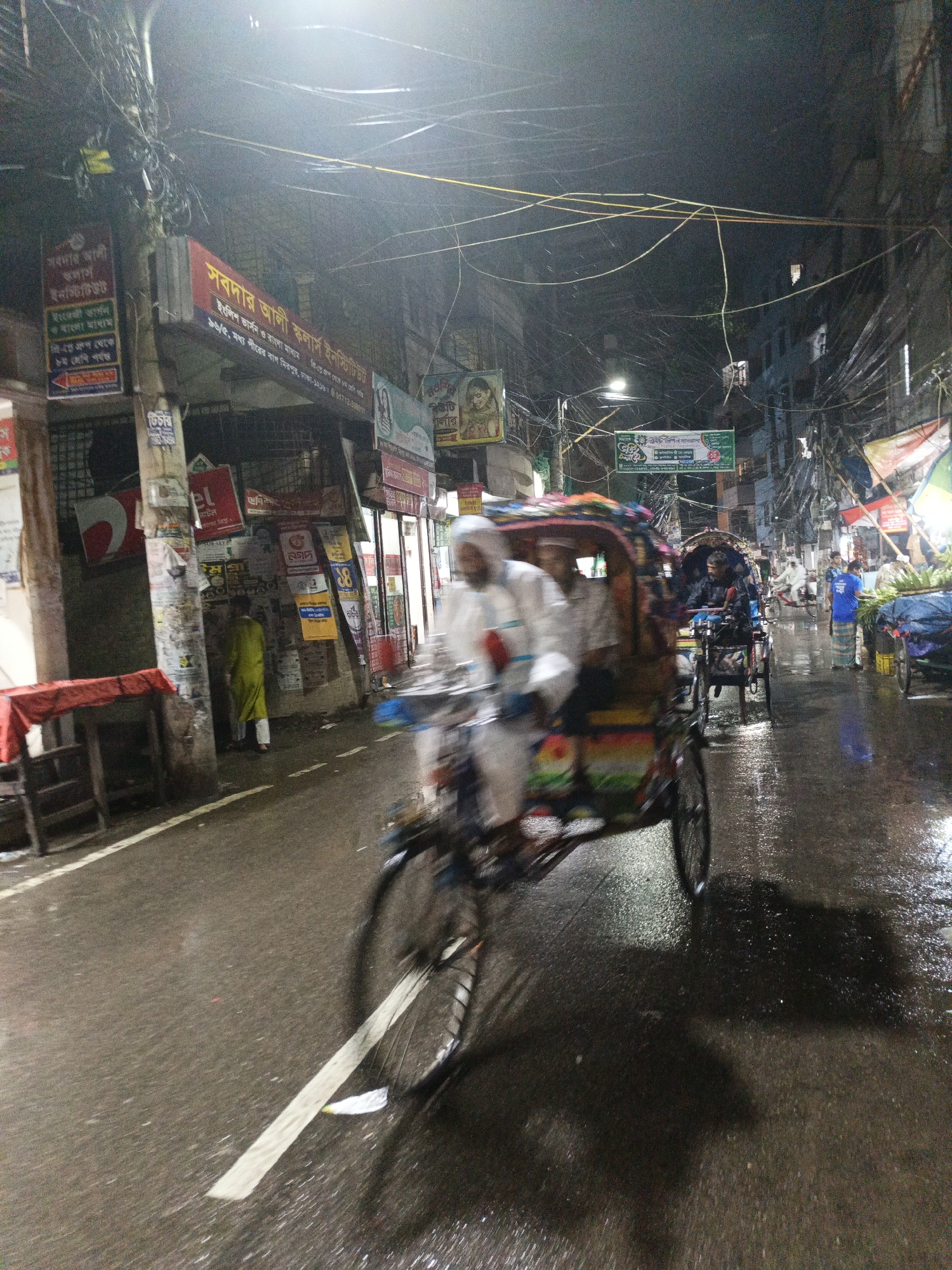 The image shows a street scene at night, likely after rainfall, as the pavement appears wet and reflective. The scene is busy with several rickshaws moving along the street. One rickshaw is prominently blurred in the foreground, indicating its movement. The passenger(s) are visible inside. Further down the street, another rickshaw with a person under an umbrella can be seen.
On the left side of the street, there are buildings adorned with numerous advertisements and signage in Bengali script. Power lines and cables crisscross overhead, creating a dense web against the night sky. A person in a light green outfit is standing by a building column. In the background, there is another rickshaw with a vendor selling vegetables.