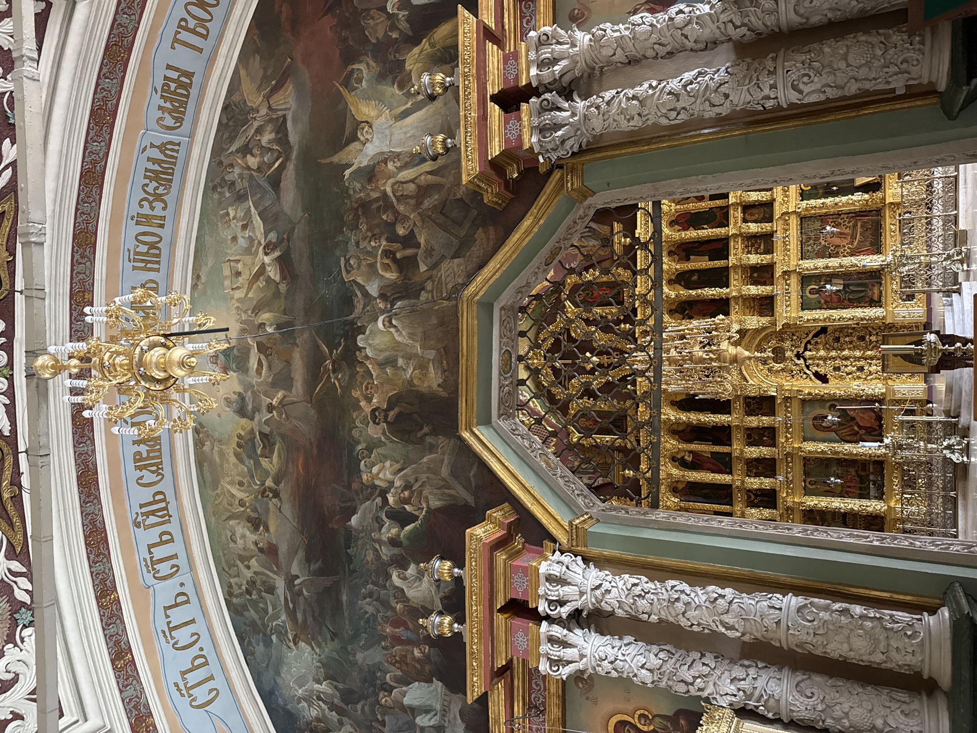 The image shows the interior of the Trinity Lavra of St. Sergius in Sergiev Posad, Russia, specifically the iconostasis in the Assumption Cathedral.
In the foreground, ornate golden columns with intricate white carvings flank a view of the iconostasis. The iconostasis is a richly decorated wall of icons, primarily in gold frames, separating the nave from the sanctuary. Several large icons are visible, depicting religious figures in traditional Byzantine style. Smaller icons are arranged in multiple tiers, creating a densely packed and visually striking display. A gold-colored metal railing runs along the base of the iconostasis.
Above the iconostasis and filling the upper part of the image is a large fresco depicting a biblical scene, likely the Last Judgment or the Ascension, with numerous figures, including angels and people, rendered in a dramatic and dynamic style. A golden chandelier hangs from the ceiling, partially obscuring the fresco. Decorative painted moldings and architectural details adorn the ceiling and upper walls.
The overall impression is one of opulence, religious grandeur, and historical significance.