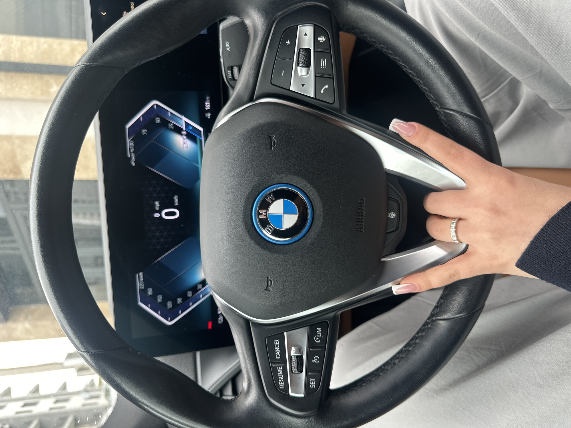 The image displays a close-up, first-person perspective from the driver's seat of a modern vehicle, focusing on the steering wheel and digital instrument cluster.
The central subject is a black leather multi-function steering wheel. Its hub features a BMW logo with a prominent blue outer ring, signifying an electric or hybrid model, and the word "AIRBAG" beneath it. The left spoke contains buttons labeled "RESUME," "CANCEL," "SET," and two additional control icons. The right spoke includes controls for audio volume (+/-), menu navigation (scroll wheel and arrows), phone, menu, and voice commands. A heated steering wheel icon is visible on the bottom right spoke.
Behind the steering wheel is a wide, digital instrument cluster displaying "0 mph" and "0 km/h" on the left, indicating the vehicle is stationary. The right side shows "ePower % 100" and "READY 0," with a visible electric range of "161 km." A red warning indicator light is active below the left gauge.
A human right hand with manicured French-tipped nails and a silver-colored ring adorned with multiple clear stones on the ring finger rests on the lower right spoke of the steering wheel. The hand's wrist is covered by a dark blue sleeve, and light grey fabric, consistent with clothing, is visible below the steering wheel.
Through the windshield, the blurred background consists of light-colored, multi-story buildings, typical of an urban setting. No specific landmarks or identifying street features are discernible to precisely locate the scene within Yerevan, Armenia.