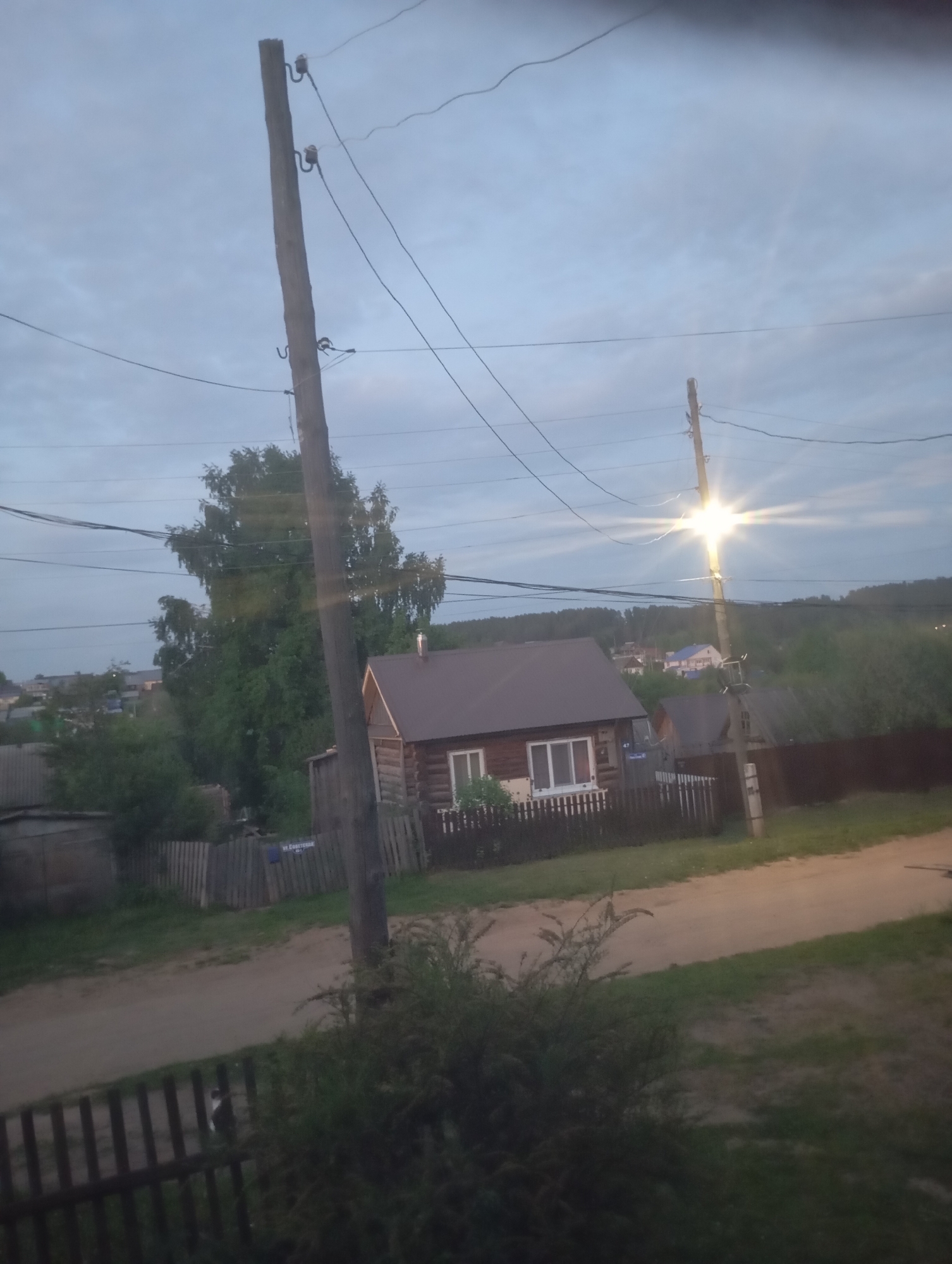 The image depicts a rural or suburban scene, possibly in a Russian village, based on the architectural style of the houses. The sky is overcast, suggesting early morning or late evening lighting conditions. Two utility poles are visible, with power lines crisscrossing the view. A wooden house with a dark roof and white-framed windows is located in the middle ground, behind a wooden fence. Another house is seen to the right. There is a dirt road running through the scene. A small bush or shrub is located in the foreground. The location is the city of Unknown, Unknown.