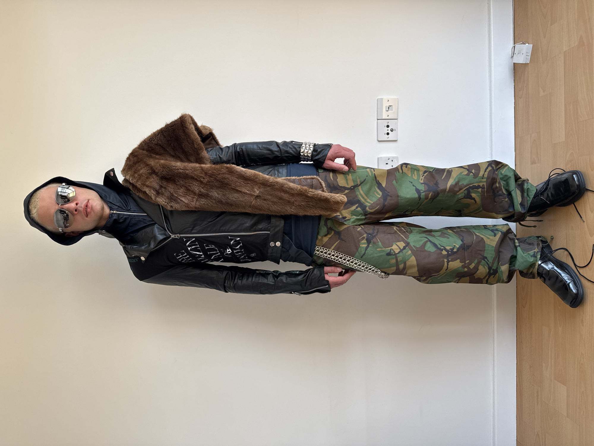 This full-length portrait, oriented with the subject standing upright, captures an individual posing indoors against a plain, light-colored wall and a light-toned wooden floor.
The subject, appearing to be an adult with short blonde hair, wears dark, mirrored sunglasses and a black hooded jacket, likely leather, featuring visible white text on the chest which appears to be part of a brand name, possibly "PLEIN DENIM." A substantial brown faux fur stole or scarf is draped over their left shoulder and across the front of their body. Underneath, a dark-colored top is visible. The person also wears a prominent silver-studded bracelet on their left wrist.
For the lower half, they are dressed in loose-fitting, camouflage-print trousers in typical green, brown, and black patterns. A silver-colored multi-chain wallet chain hangs from the right side of the trousers. The outfit is completed with shiny black lace-up shoes or boots. The subject's right hand is casually placed near their hip.
In the background, on the white wall to the right, several white electrical outlets and sockets are visible, consistent with UK standards, providing a location cue for London, United Kingdom. The overall lighting is even, suggesting an indoor setting. The subject's face is partially obscured by the sunglasses, displaying a neutral expression as they pose.