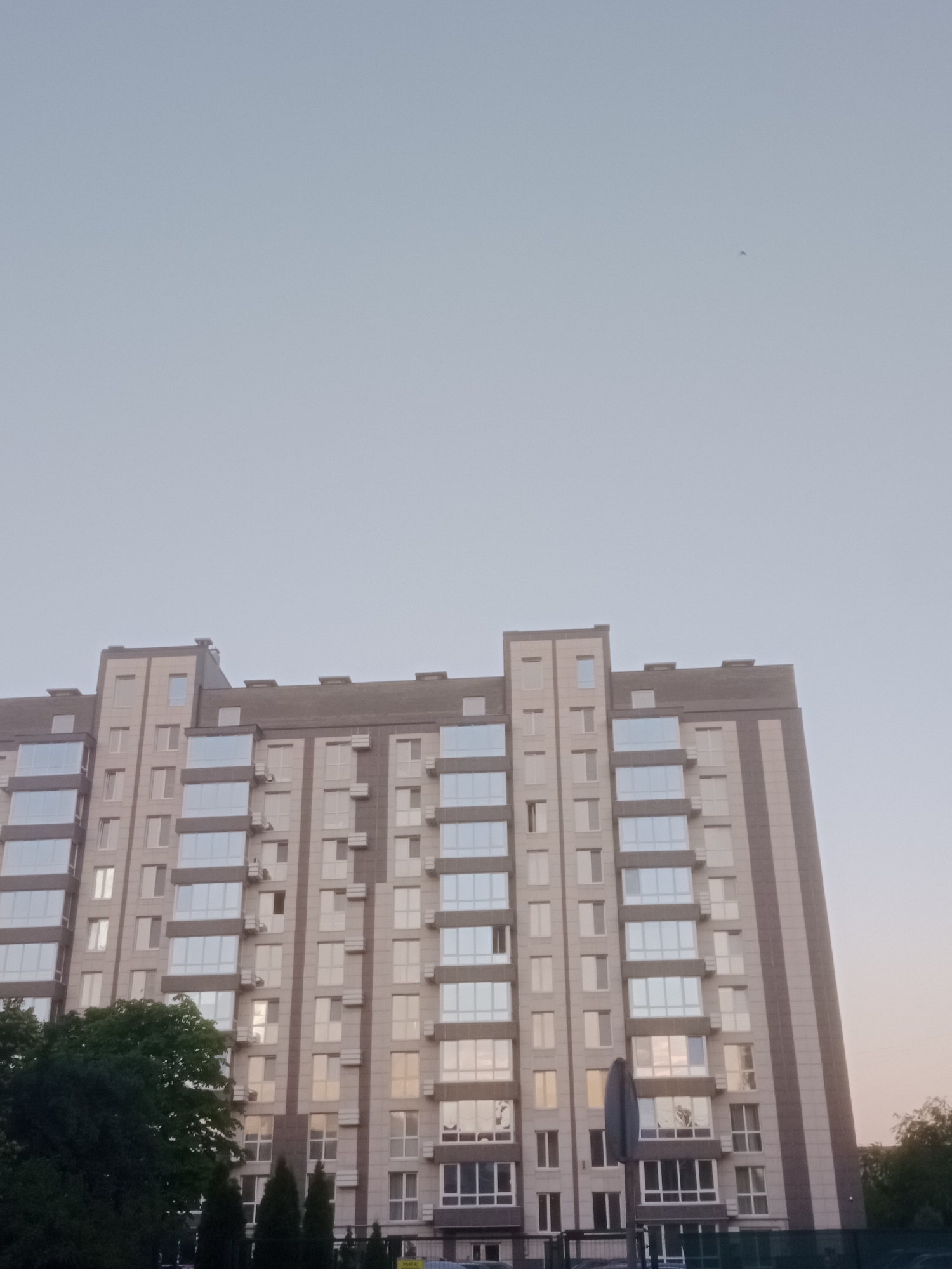 The image shows a tall apartment building against a clear, pale blue sky. The building has a modern design with a facade of light-colored tiles and numerous windows. The windows are reflective, showing the sky and a hint of sunset colors. There are also some darker vertical elements on the building's facade.
In the lower left of the image, a dense green tree is visible. In front of the building, there are some smaller, dark green coniferous trees and a street sign. The overall impression is of a residential area in the city of Horishni Plavni, Ukraine, possibly captured during the late afternoon or early evening. The sky is mostly clear, with a very faint speck visible in the upper right quadrant, which could be a bird or a distant aircraft. There are no people visible in the image.