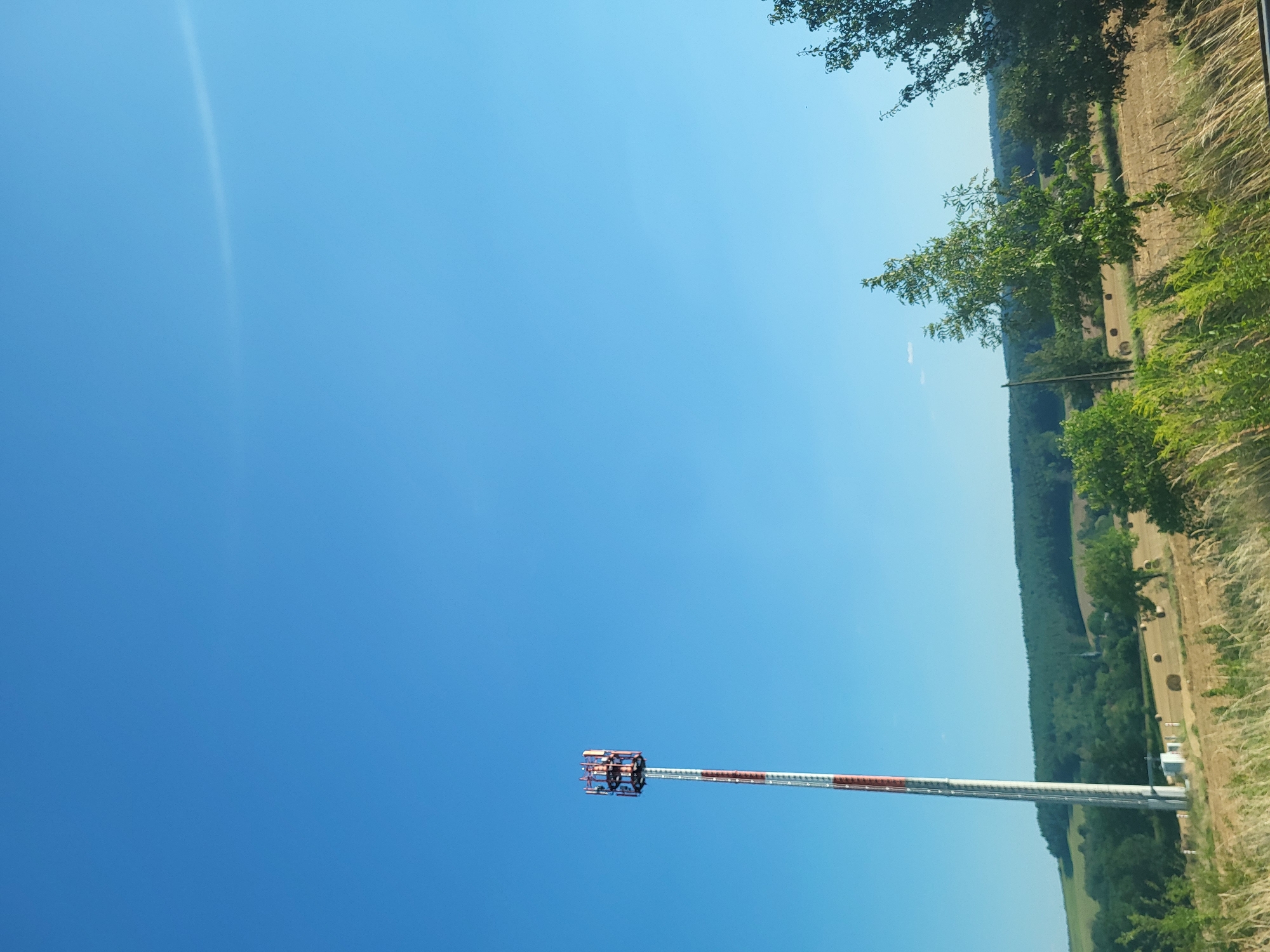 This media file captures a clear, bright day with a vast expanse of blue sky dominating the frame. In the foreground, a tall, slender tower with red and white banding extends diagonally upwards. At its apex, a complex structure with multiple levels and visible metal components suggests it's a telecommunications tower or similar infrastructure.
The background reveals a pastoral landscape, indicative of a rural setting. A sloping field, likely agricultural land, is dotted with what appear to be round hay bales. Beyond this, a dense line of green trees forms a natural barrier, suggesting a forest or wooded area. The edge of the frame on the right side shows more vegetation, possibly grasses or hedges, and hints at a roadside or field boundary.
The lighting and the clear blue sky indicate a time of day likely late morning or early afternoon, with sunny weather. The scene evokes a sense of openness and tranquility, common to rural German landscapes. There is no visible text in the image. The orientation of the image is such that the tower appears to be in the lower right quadrant, and the landscape is visible in the upper right quadrant.