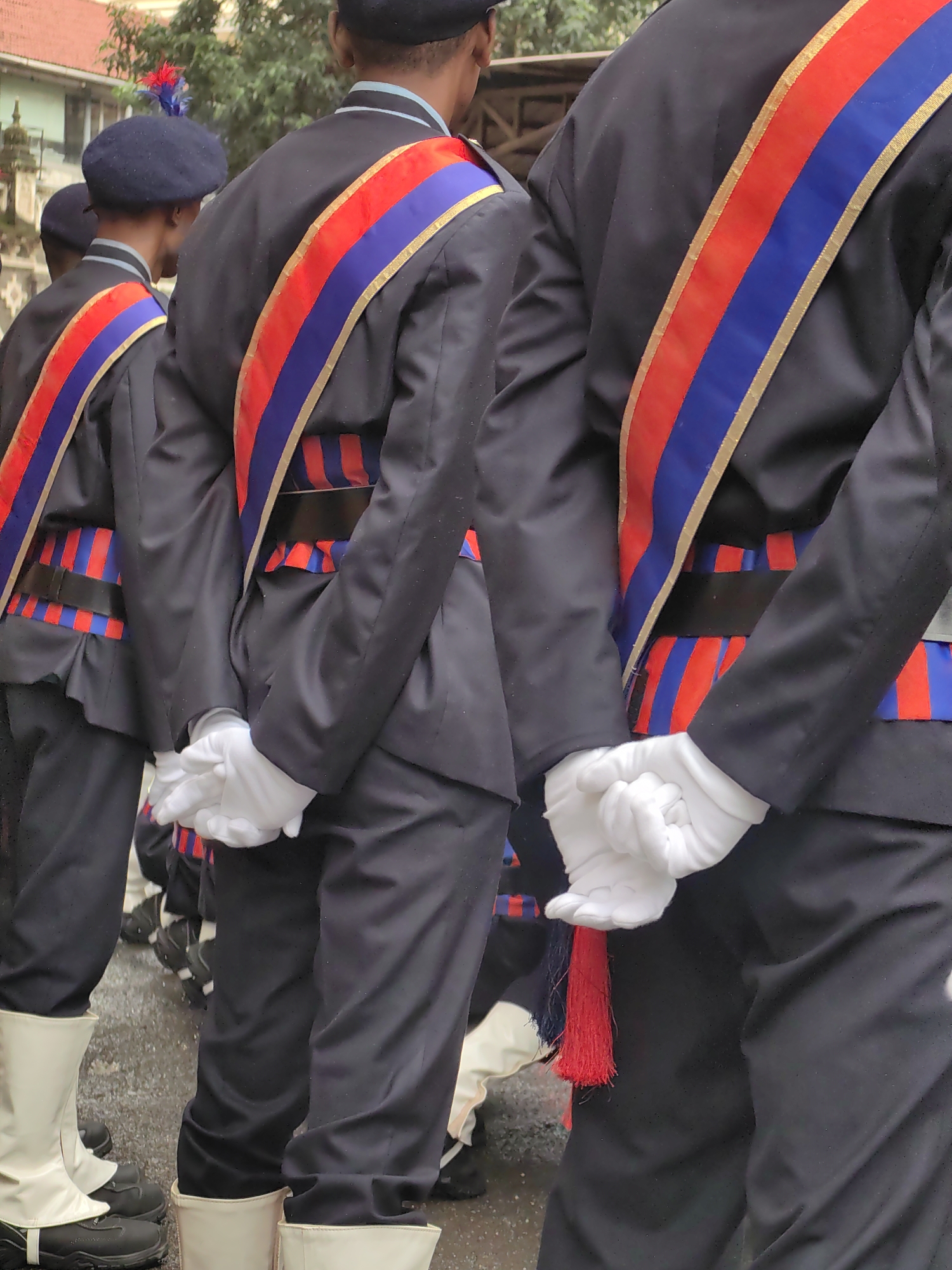 The image depicts a group of individuals in ceremonial attire, standing in what appears to be a city street. The individuals are wearing dark uniforms with prominent red, blue, and gold sashes across their chests. Some are also wearing white gloves and white gaiters over dark footwear. The background shows some buildings and foliage, suggesting an outdoor urban setting. The scene is likely a parade or official ceremony. The specific location in Mumbai, India, cannot be determined from the visual information alone.