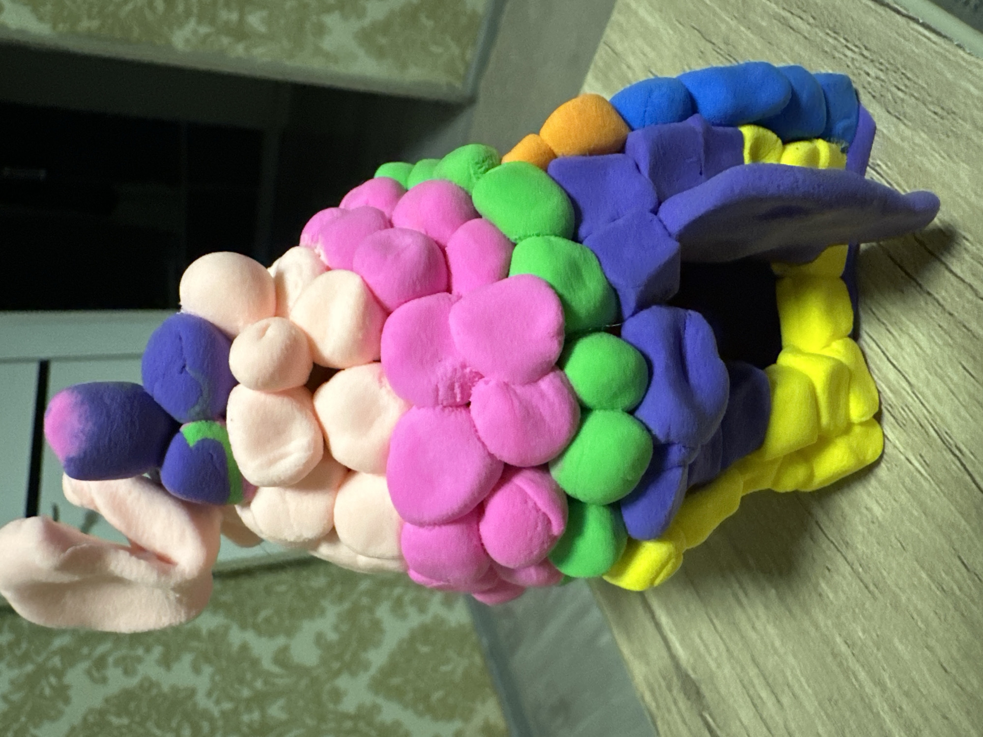A brightly colored, homemade sculpture crafted from modeling clay sits on a wooden table. The sculpture, which resembles a layered, whimsical tower, is composed of numerous small, rounded pieces of clay arranged in concentric circles and stacked vertically. The colors used include pink, light pink, purple, green, yellow, orange, and blue. The sculpture has an organic, somewhat abstract form with a light pink, ear-like protrusion extending from the top left. The background consists of a wall with a patterned wallpaper and a dark doorway. The lighting suggests an indoor setting. The origin of this image is not known, and it is not possible to determine its location in Fergana City, Uzbekistan.