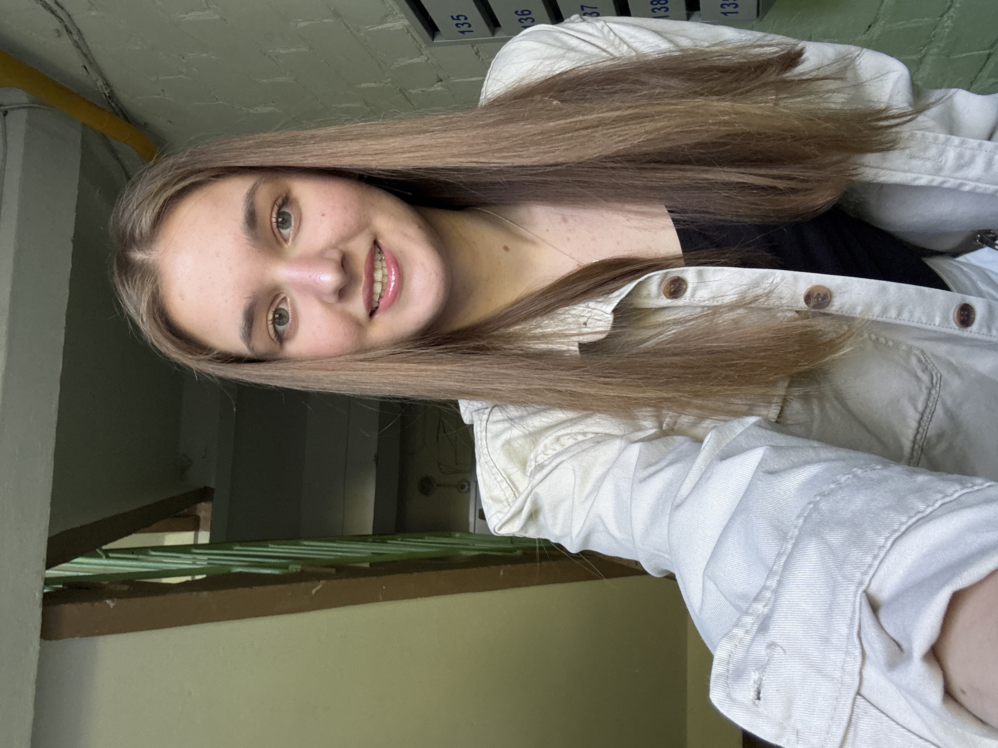 A young woman with long brown hair is taking a selfie indoors in what appears to be a stairwell. She is wearing a white button-down jacket over a black shirt. Behind her are brick walls with mailboxes labeled with numbers in the 130s. Part of a metal railing and stairwell wall are also visible. Based on the appearance of the stairwell, the presence of mailboxes and information from a search query, this is likely a residential building in Perm, Russia.