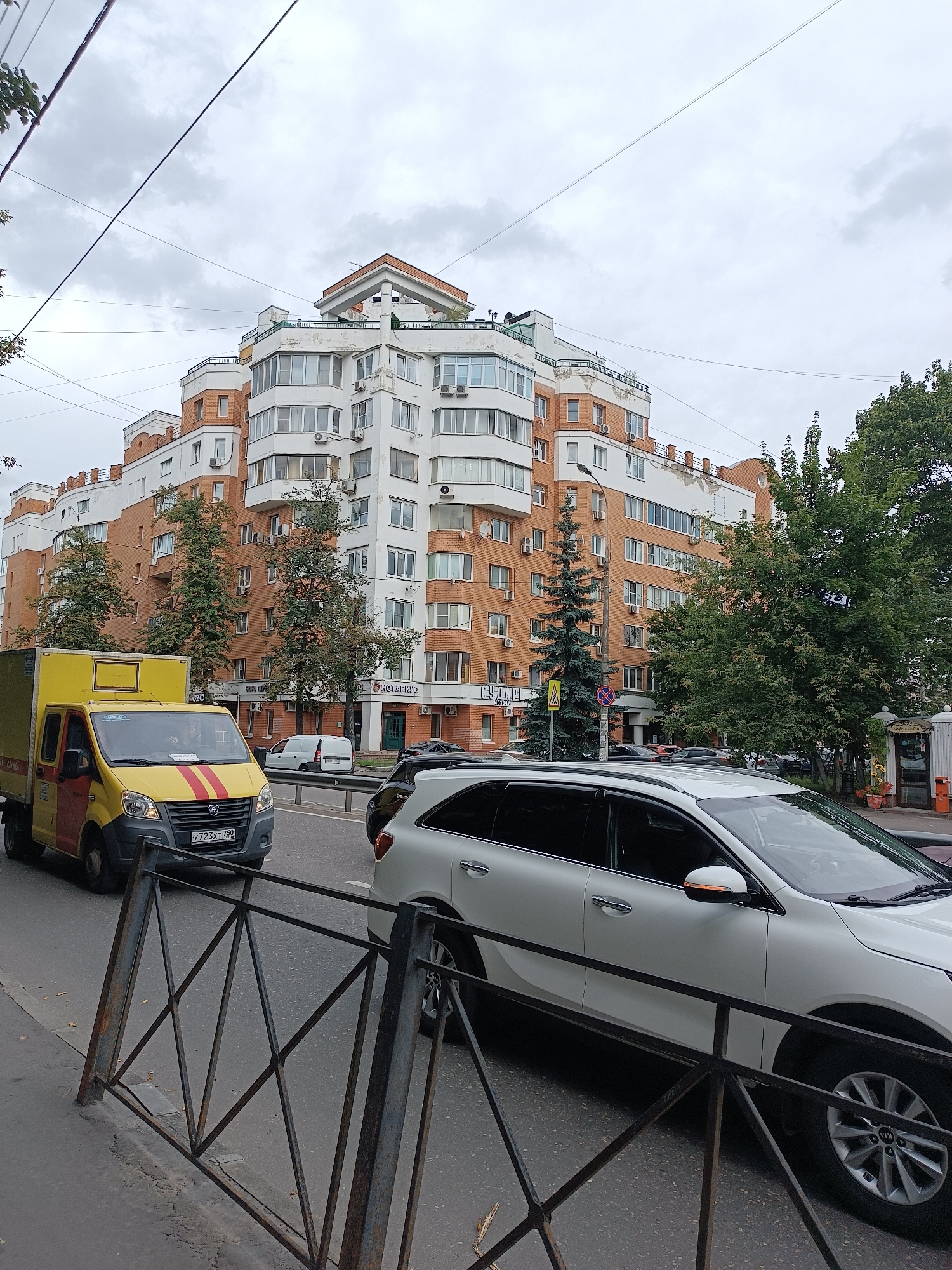 In Lyubertsy, Russia, on a cloudy day, a white Kia SUV is parked on the right side of the road, facing forward. To its left, a yellow and red delivery truck with red stripes on its hood is driving on the road. In the background, a large, multi-story building with orange brick and white sections stands tall, featuring numerous windows and balconies. Trees line the street in front of the building. A metal fence runs along the sidewalk in the foreground.