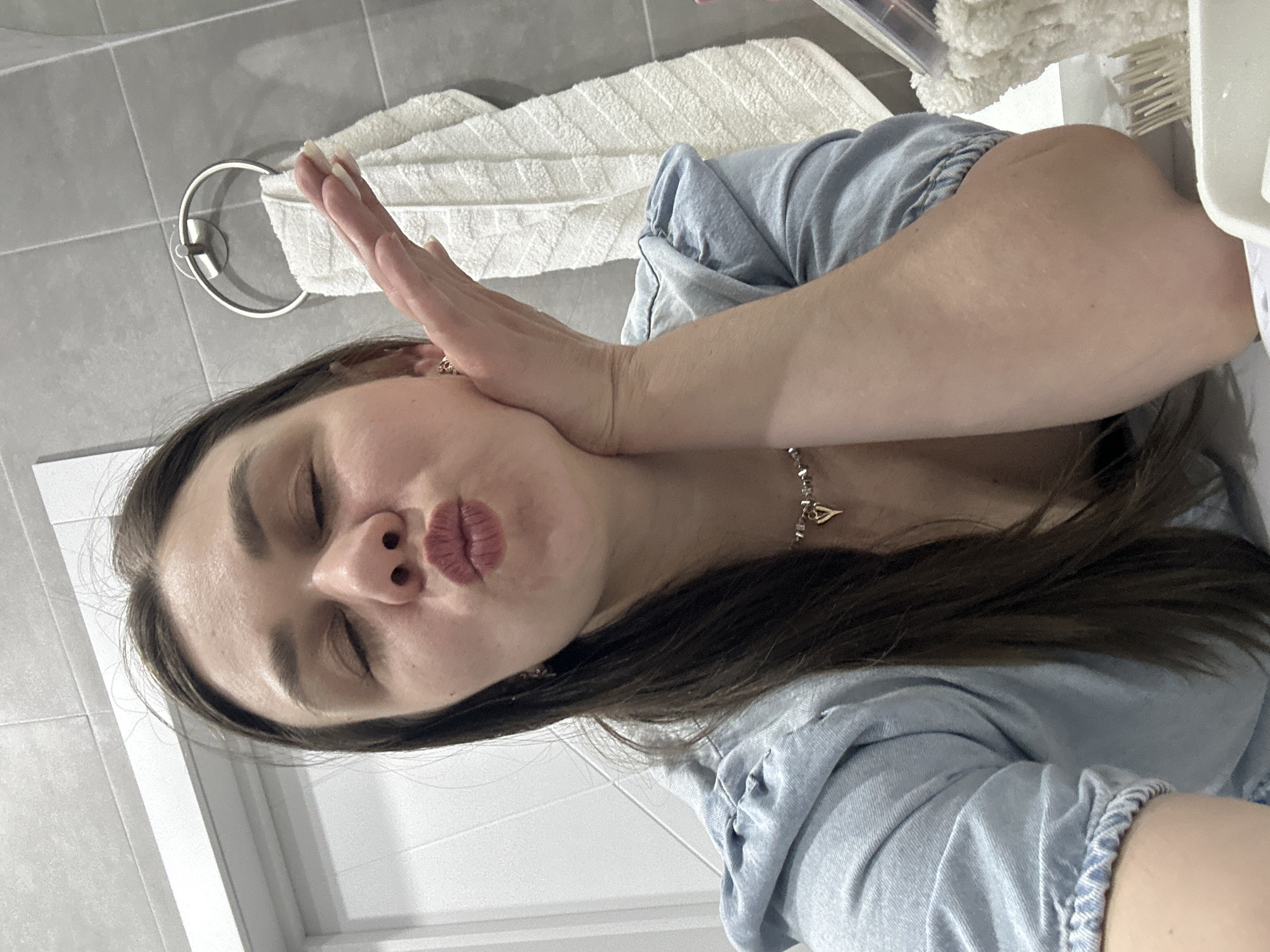 A woman with long, dark hair is posing for a selfie in a bathroom. Her eyes are closed, and her lips are puckered into a kissing expression. Her left hand is resting on her cheek. She is wearing a light blue denim shirt and a silver necklace. Behind her, a white towel is draped over a towel rack on a gray-tiled wall. The location is a bathroom, possibly in Chaikovsky, Russia, but without further context or specific details identifying the city, it is impossible to confirm.