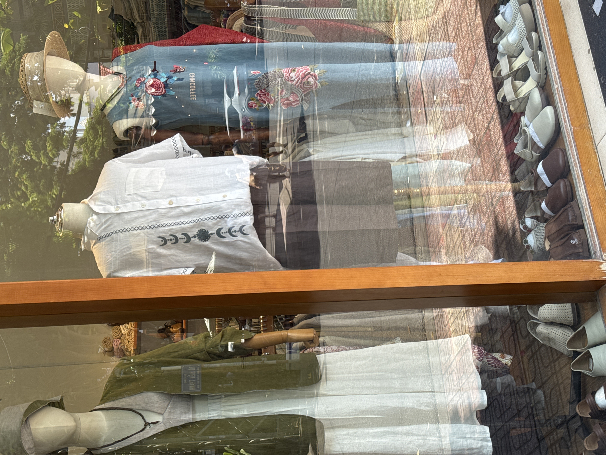 This image depicts a storefront window in Nha Trang, Vietnam, showcasing a display of clothing and accessories. The display features three mannequins. The leftmost mannequin wears a light-colored dress with a green jacket and a woven hat. The central mannequin is dressed in a white short-sleeved shirt with embroidered details. The rightmost mannequin sports a blue garment with floral embroidery and a straw hat. Behind the mannequins, various folded fabrics and clothing items are visible. The floor inside the store has a tiled pattern. Through the window, reflections of the street outside can be seen, including trees, buildings, and a white car. A rack of handbags and scarves is also partially visible. In the foreground, along the bottom of the window, a collection of shoes is displayed on a shelf. The overall scene captures a retail environment, likely a boutique or clothing store.