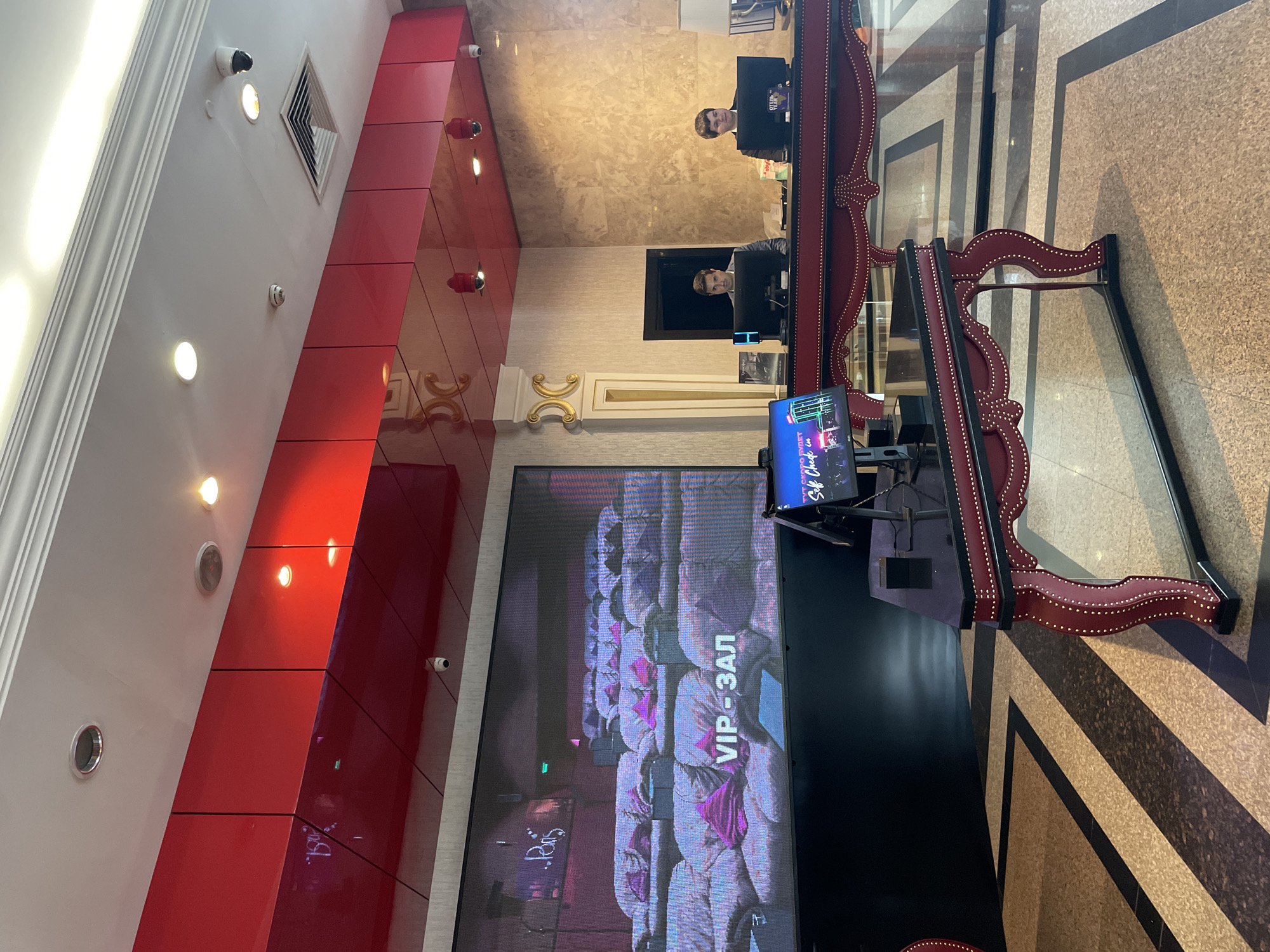 In an indoor, well-lit lobby, likely a hotel or entertainment venue, a large digital screen displays a seating area with plush sofas. Text overlayed on the screen reads "VIP-ЗАЛ", indicating a VIP lounge. Two men in formal attire are seen behind a reception desk, each attending to a computer monitor. The reception desk is ornate, with a dark red velvet-like material studded with gold accents. The floor features a geometric pattern of beige and black tiles. Red panels with embedded lights form a decorative ceiling element. The overall ambiance suggests a high-end establishment. Based on the presence of Russian text on the screen and a sign visible on one of the computers which reads "СКОРО БУДЕТ SELF CHECK IN", this scene is likely located in Kazan, Russia, a city known for its entertainment venues.