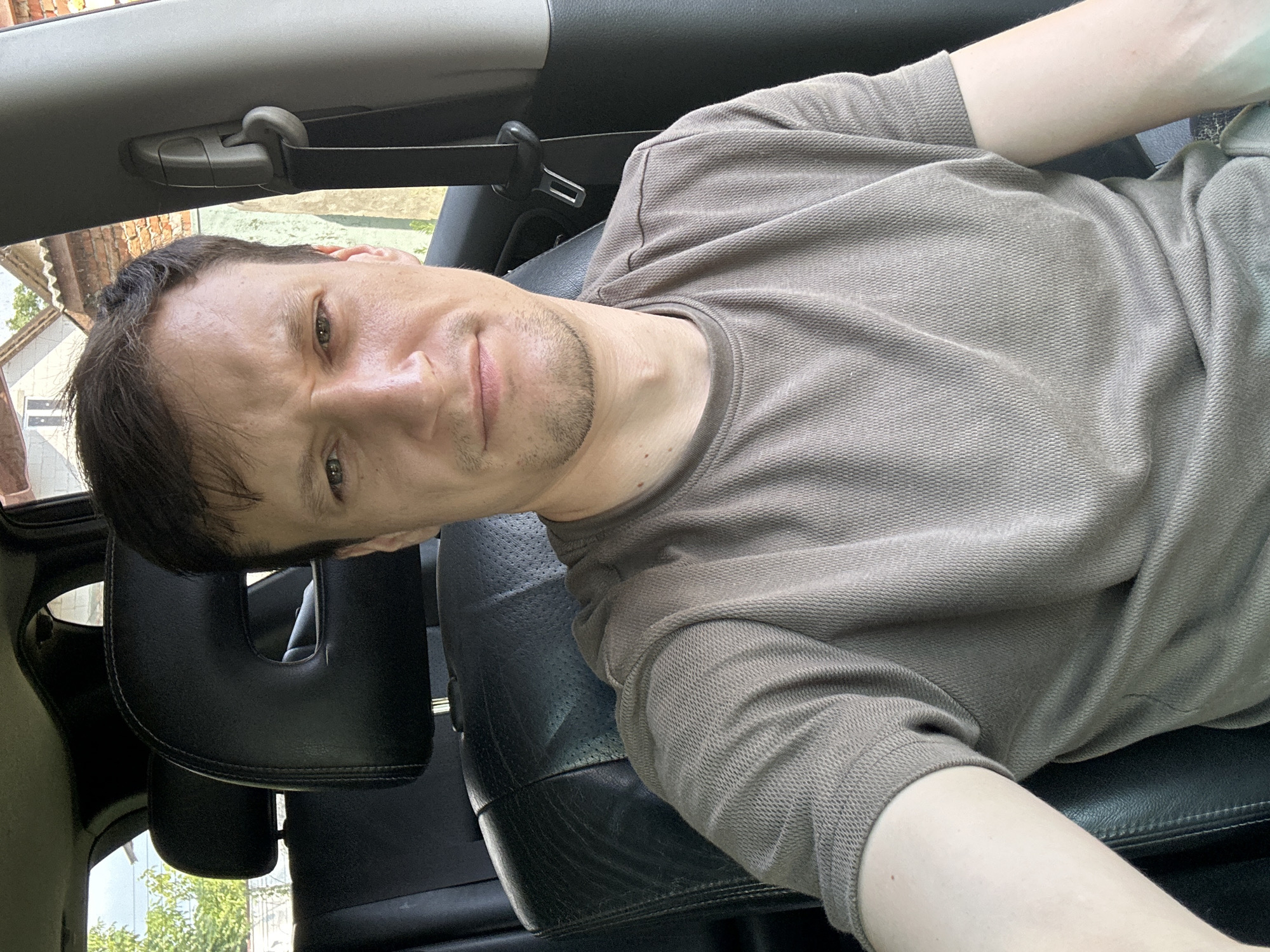 A middle-aged man with short, dark hair and a slight stubble is captured in a selfie taken from inside a vehicle. He is seated in the driver's seat, facing the camera with a neutral expression. The man is wearing a short-sleeved, taupe t-shirt. The interior of the car features black leather seats and headrests. A grey seatbelt is visible to the right of the man. Through the car windows, a residential area is visible, with buildings constructed of brick and white siding, as well as trees and foliage. The scene appears to be in Makiv, Ukraine.
