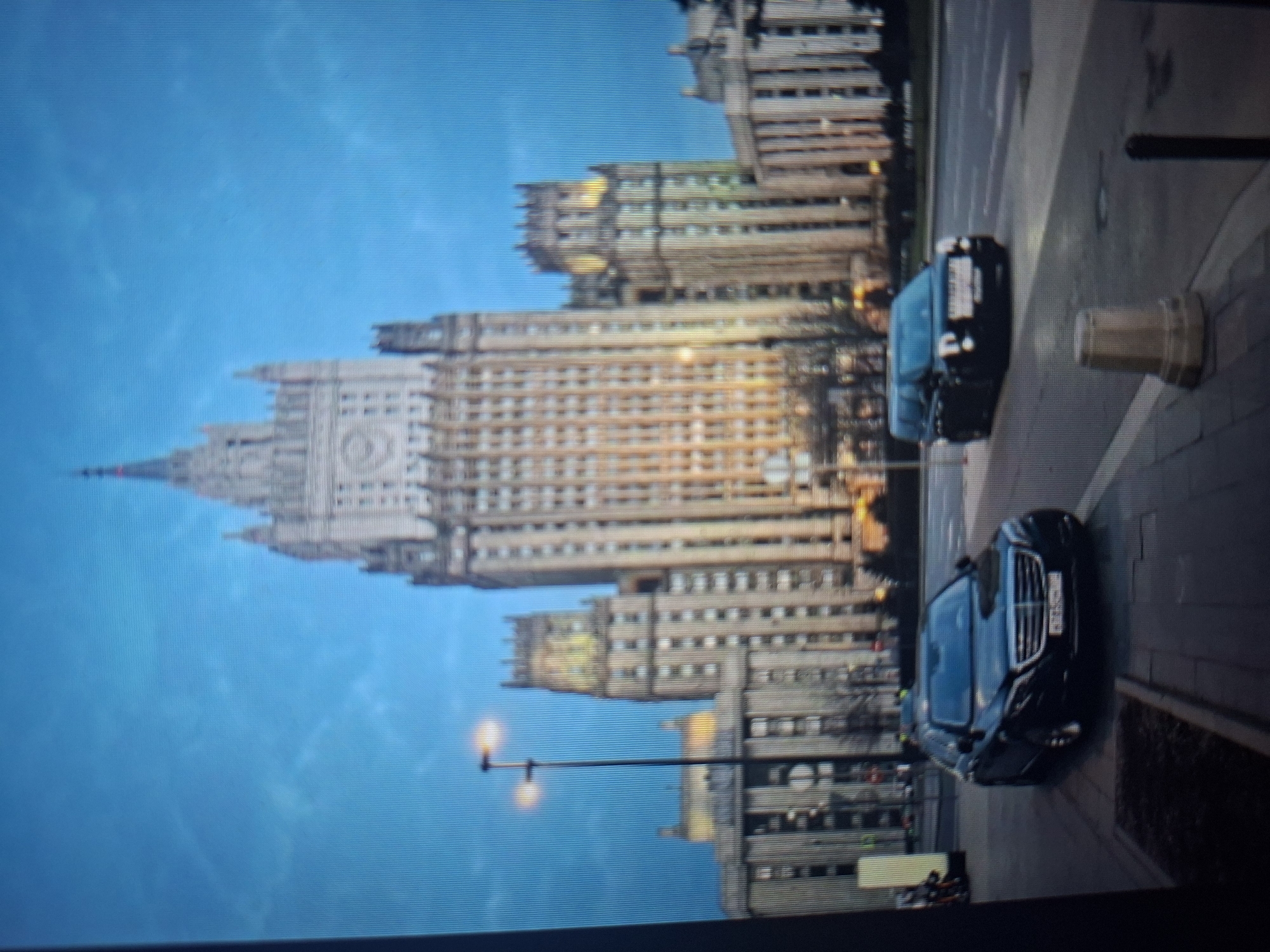 The image depicts a twilight scene in a city, featuring a prominent Stalinist skyscraper in the background. The skyscraper, likely a government building due to its style and imposing architecture, is illuminated, with visible windows and decorative elements. In the foreground, a black Mercedes-Benz sedan is parked on the right side of the street, facing forward. To the right of the sedan, a black van is driving on the road. A street lamp with two lights is visible to the left of the skyscraper, casting an orange glow. The sky is a deep blue, suggesting either dawn or dusk, with some faint clouds. The area appears to be a public space with a sidewalk and a street. There is no indication that this scene is located in Užuežerė, Lithuania; the architecture is characteristic of Moscow or other cities in former Soviet republics.