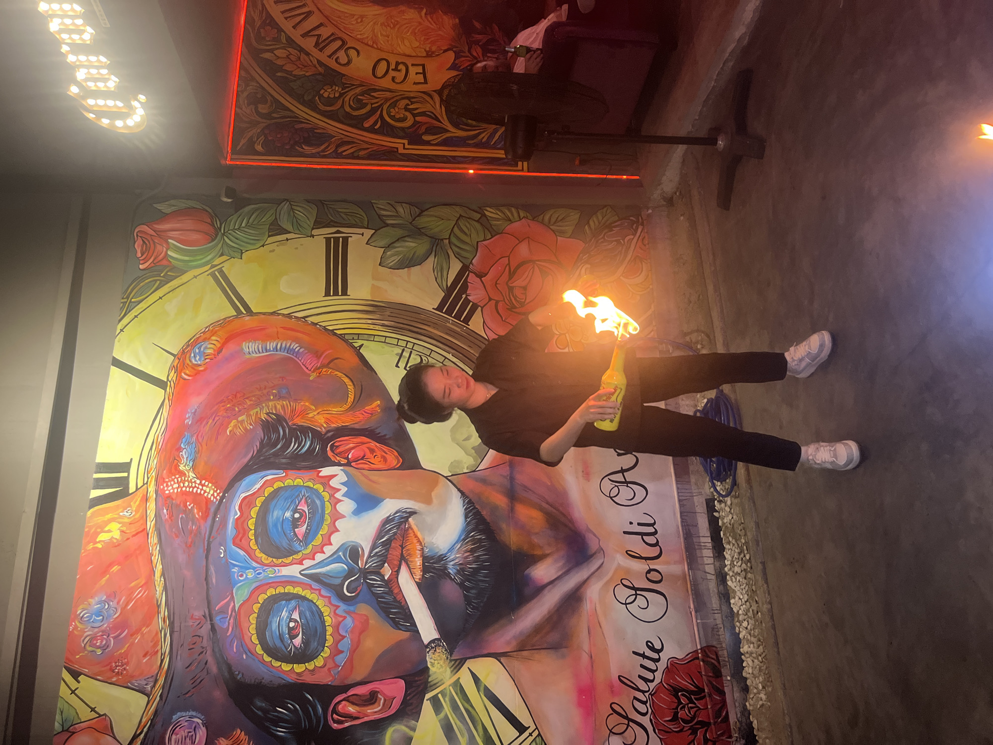 The image captures a dimly lit indoor space in Da Nang, Vietnam, likely a bar or restaurant, adorned with vibrant murals. Dominating the background is a large, colorful mural depicting a stylized skull with a wide-brimmed hat, reminiscent of Dia de los Muertos imagery. The skull is rendered with bold colors and intricate patterns. To the right of the mural, a neon sign that reads "Corona" is visible.
In the foreground, a young Asian woman, dressed in a black collared shirt and black pants, stands facing the viewer. She is holding a bright yellow bottle, from which flames are erupting, suggesting she is performing a fire-eating or fire-based act. Her stance is casual, with her left leg slightly forward, and she is looking down at the bottle with a focused expression. She is wearing white sneakers.
The environment appears to be a tiled floor with scattered gravel. A black floor fan is positioned to the right of the woman. In the mid-ground, partially obscured, a person is seated on a couch, holding a bottle. The overall atmosphere is one of entertainment and vibrant cultural display, typical of a lively establishment in Da Nang.