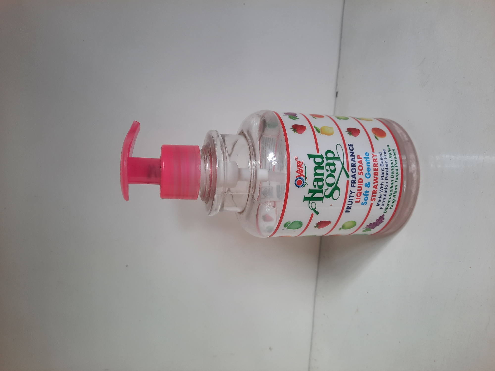 A clear plastic bottle of YURI brand Strawberry scented liquid hand soap with a pink pump dispenser sits on a white surface. The bottle has a label with colorful fruit illustrations and text that reads "YURI Hand Soap Fruity Fragrance Liquid Soap Soft & Gentle STRAWBERRY Made With Plant Based Formulation Paraben Free Di formulasikan Dengan Bahan Alami Tanpa Paraben". The background is a plain white wall. The scene is indoors, with no indication of location in Lhokseumawe, Indonesia.