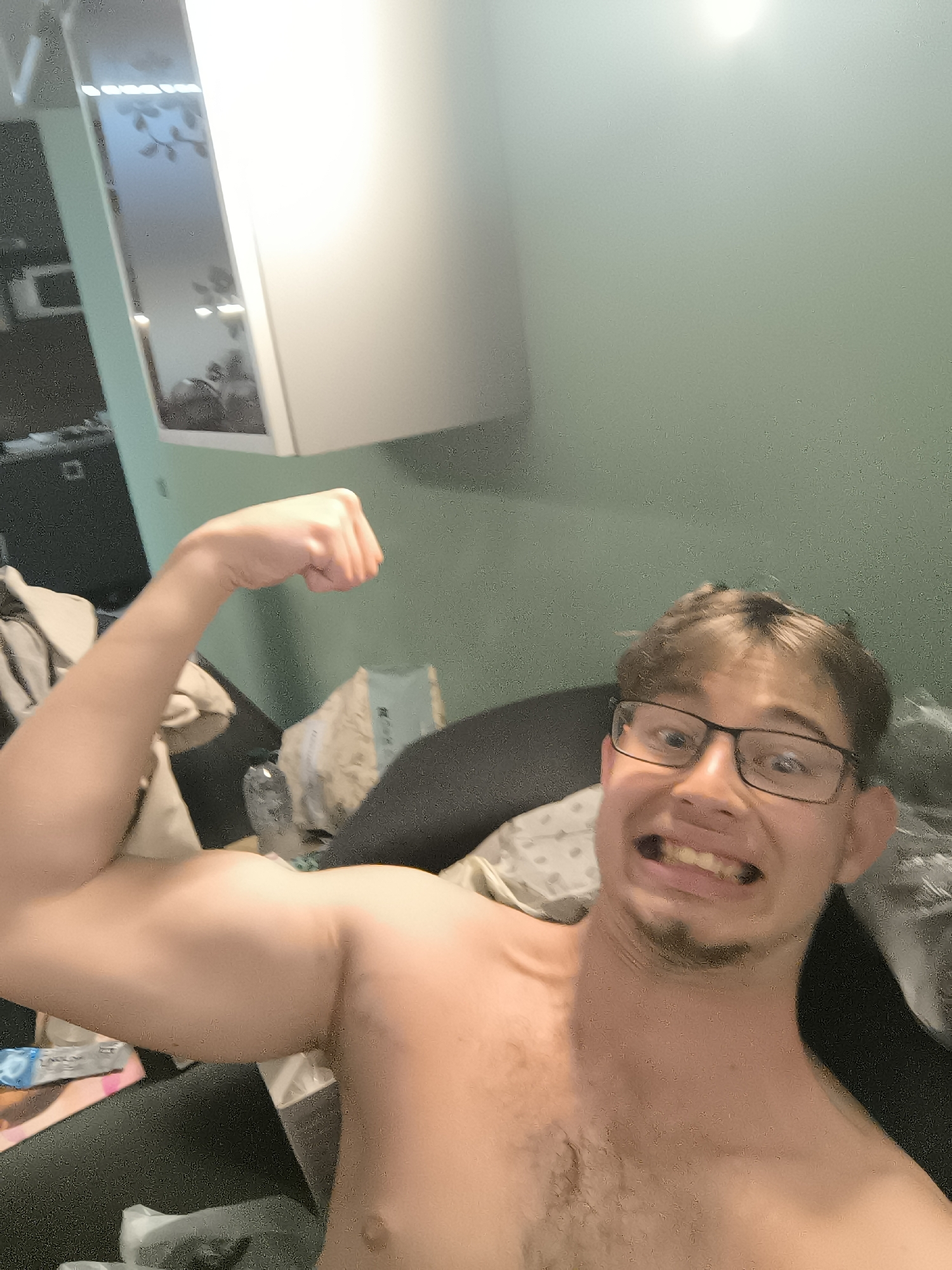 A shirtless young man wearing glasses flexes his left bicep. He is sitting on a dark-colored upholstered surface, potentially a sofa or chair. Behind him is a green wall and a white cabinet. The setting appears to be indoors, possibly an apartment. There is no verifiable information to determine the location to be Sofia, Bulgaria.
