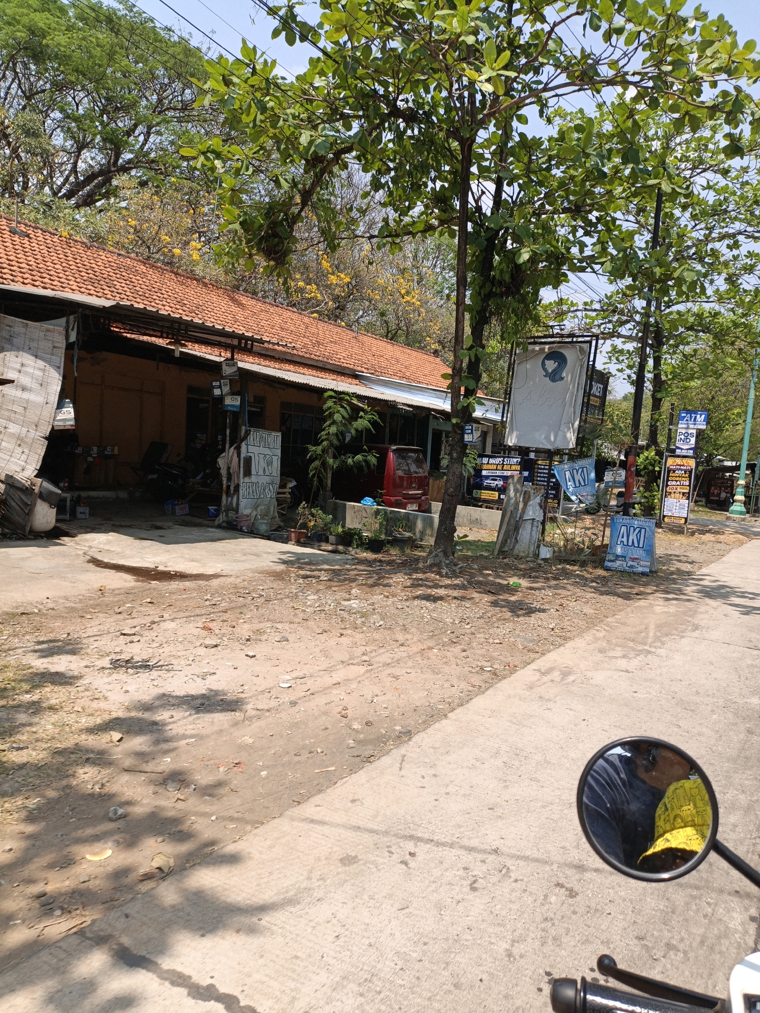 The image captures a street view in Ujungbatu, Indonesia. In the foreground, a paved road is visible with shadows cast by trees. To the left, a dirt area leads to a small building with an orange tiled roof. This building appears to be a shop or workshop, with various items, including batteries and what looks like a motorcycle, displayed or stored outside. Several signs are present, with some in Indonesian. One sign reads "TUKAH TAMBAH AKI" which translates to "Battery Exchange." There are also signs for "GS" batteries. In the background, dense green trees dominate the upper portion of the image, with some smaller trees and yellow flowers. A red van is parked in front of the building. In the reflection of a motorcycle mirror on the right side of the frame, a person wearing a yellow head covering and a dark blue shirt is visible.