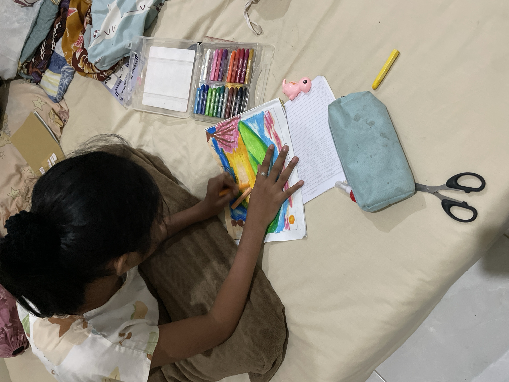 A young individual is drawing on a bed in what appears to be a residential setting in Blang Cut, Indonesia. The individual, with dark hair pulled back in a bun, is wearing a light-colored shirt with a pattern of fruit and clouds. They are focused on a drawing, holding an orange oil pastel in their right hand, actively coloring a landscape scene that includes a volcano, a body of water, and a sky with yellow and red hues.
Scattered around the drawing are art supplies, including a clear plastic case filled with oil pastels of various colors, a pink fish-shaped toy, a notebook with handwritten lines, a light blue pencil case, a pair of black-handled scissors, and a yellow oil pastel. The bed is covered with a light beige sheet, and several patterned fabrics and a book are visible in the background. The overall impression is of a child engaged in a creative activity in a relaxed, home environment.