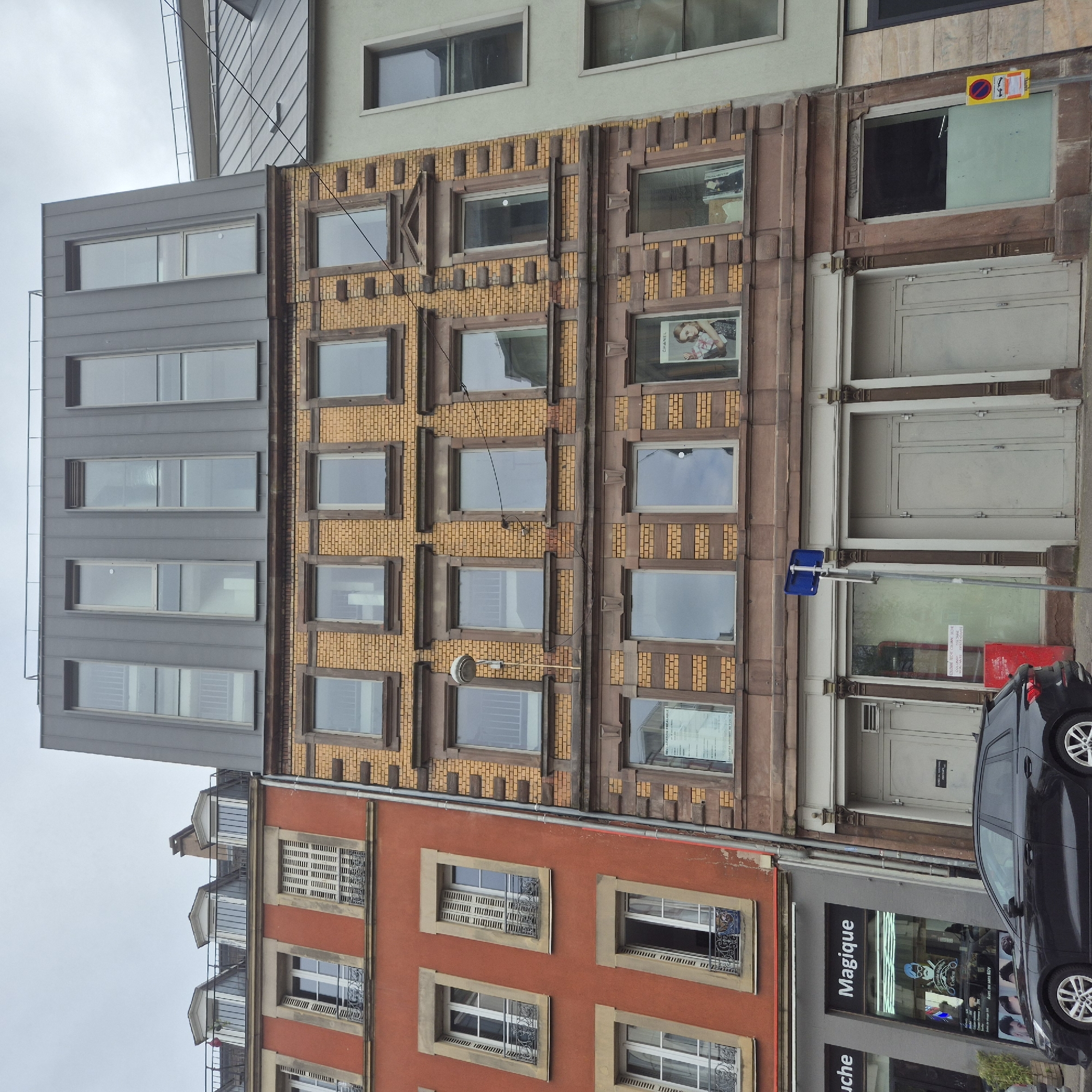 The image depicts a street scene in Strasbourg, France, showcasing a row of closely situated buildings exhibiting varied architectural styles. From left to right, the buildings are characterized by a contemporary structure with a flat, gray façade and uniform rectangular windows; a brick building featuring rows of windows outlined by ornamental brickwork; and a building with a red facade and elaborate window frames.
A black car is parked on the street, partially obscuring a storefront with "Magique" displayed on the signage. Above this business is a building with pale-colored doors and frames and a small red box.
A street sign is also evident.
The image is oriented at an angle, with the horizon tilted. The sky is overcast, and the scene is generally absent of human activity. There are signs in the windows of the buildings.