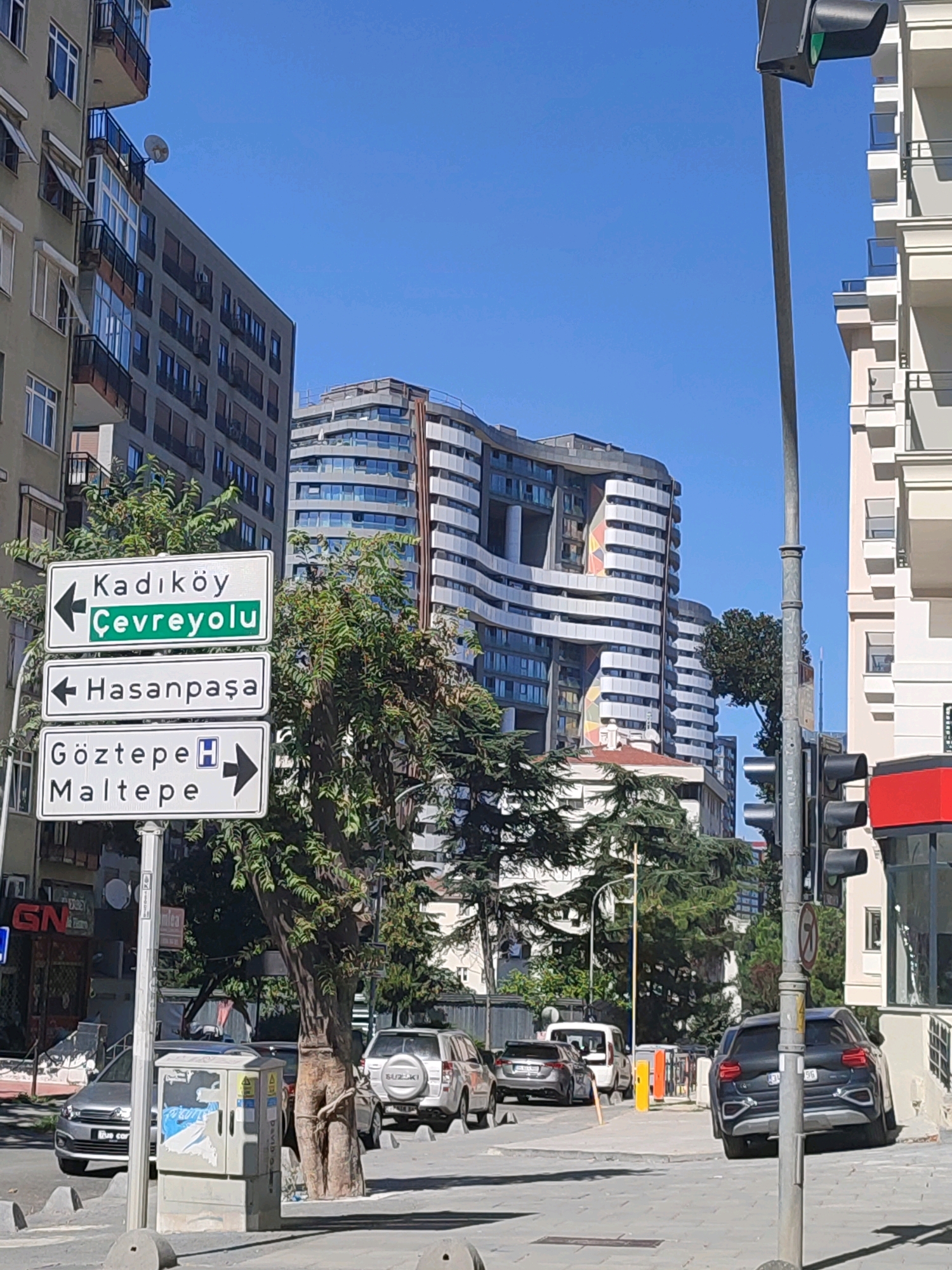 A street scene in Kadıköy, Turkey, displays a clear blue sky above modern buildings and signage. Three stacked white street signs with black lettering are prominently positioned on the left, indicating directions to "Kadıköy Çevreyolu" to the left, "Hasanpaşa" to the left, and "Göztepe Maltepe" to the right, with a hospital symbol next to Göztepe.
The environment includes a traffic light pole on the right, a sidewalk with concrete bollards, and paved road surfaces. Several cars are visible, including a silver SUV with "SUZUKI" branding on the rear, a silver sedan, a black SUV, and a white van. Buildings range in style from older, more traditional apartment blocks to contemporary structures with curved facades and numerous windows. Lush green trees and foliage are interspersed between the buildings and along the street. Some retail establishments are visible in the background. The overall scene depicts a typical urban street environment on a sunny day.