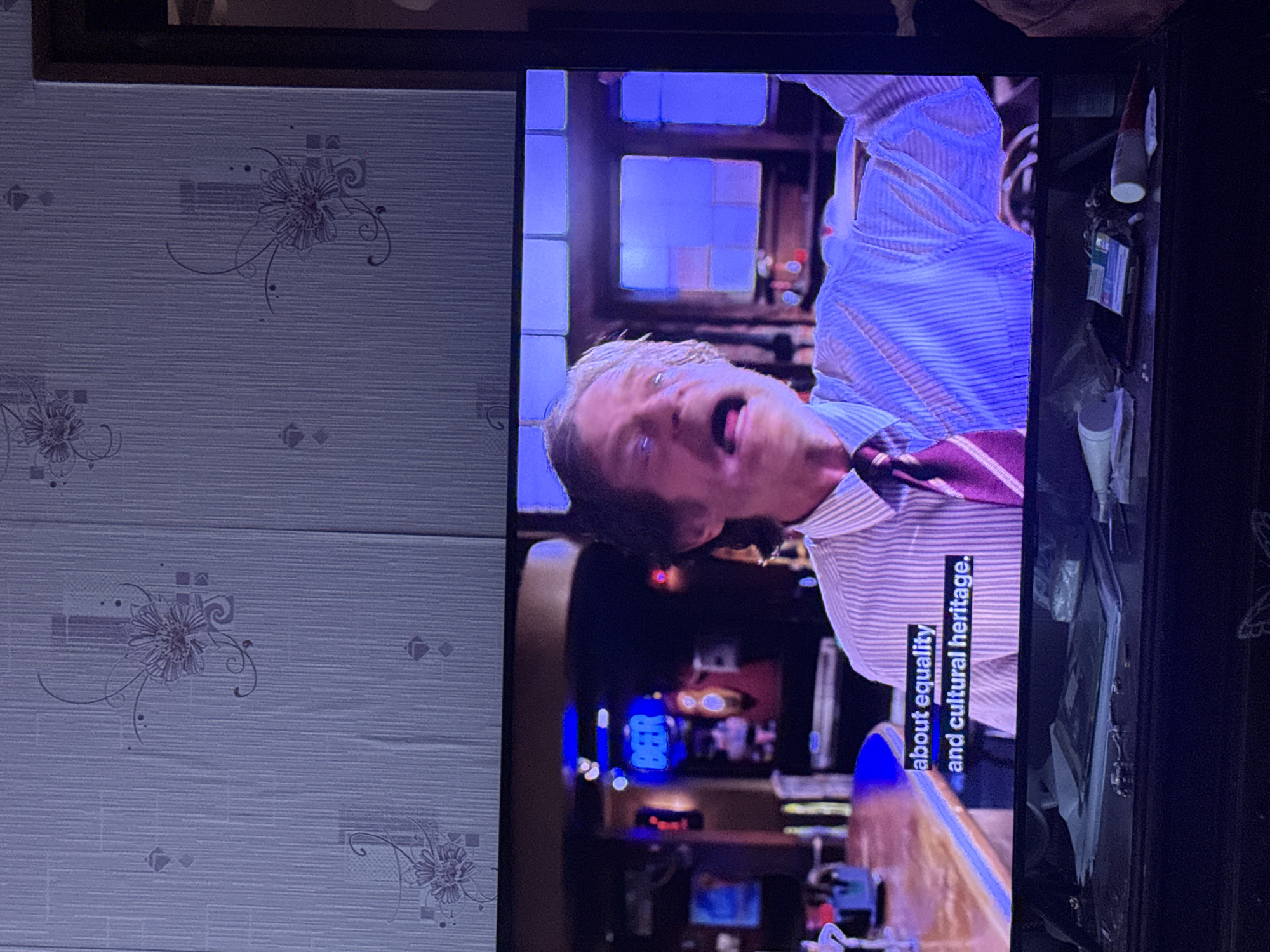 The image shows a television screen displaying a scene of a man in a striped shirt and tie, appearing to speak. Text on the screen reads "about equality and cultural heritage." The background of the scene on TV appears to be a bar setting with illuminated signs. The television is positioned next to a wall with patterned wallpaper. There is no information in the image that refers to Vratsa, Bulgaria. The description contains only details observed from the image.
