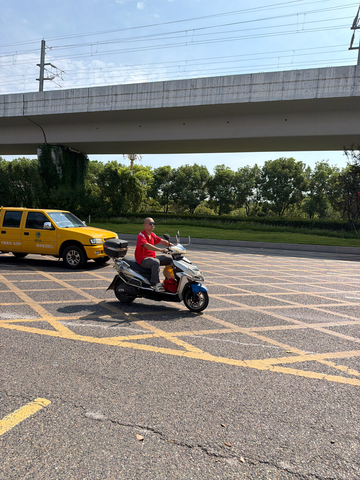 A man in a red shirt and dark pants rides a white and blue scooter on a road marked with yellow grid lines. He is wearing sunglasses. To his left, a yellow pickup truck with a camper shell is parked. The truck has Chinese text on its side. In the background, a concrete overpass with electrical wires runs overhead. Lush green trees line the side of the road. The sky is blue with scattered clouds. This scene is located in Jiangjin District, Chongqing, China.