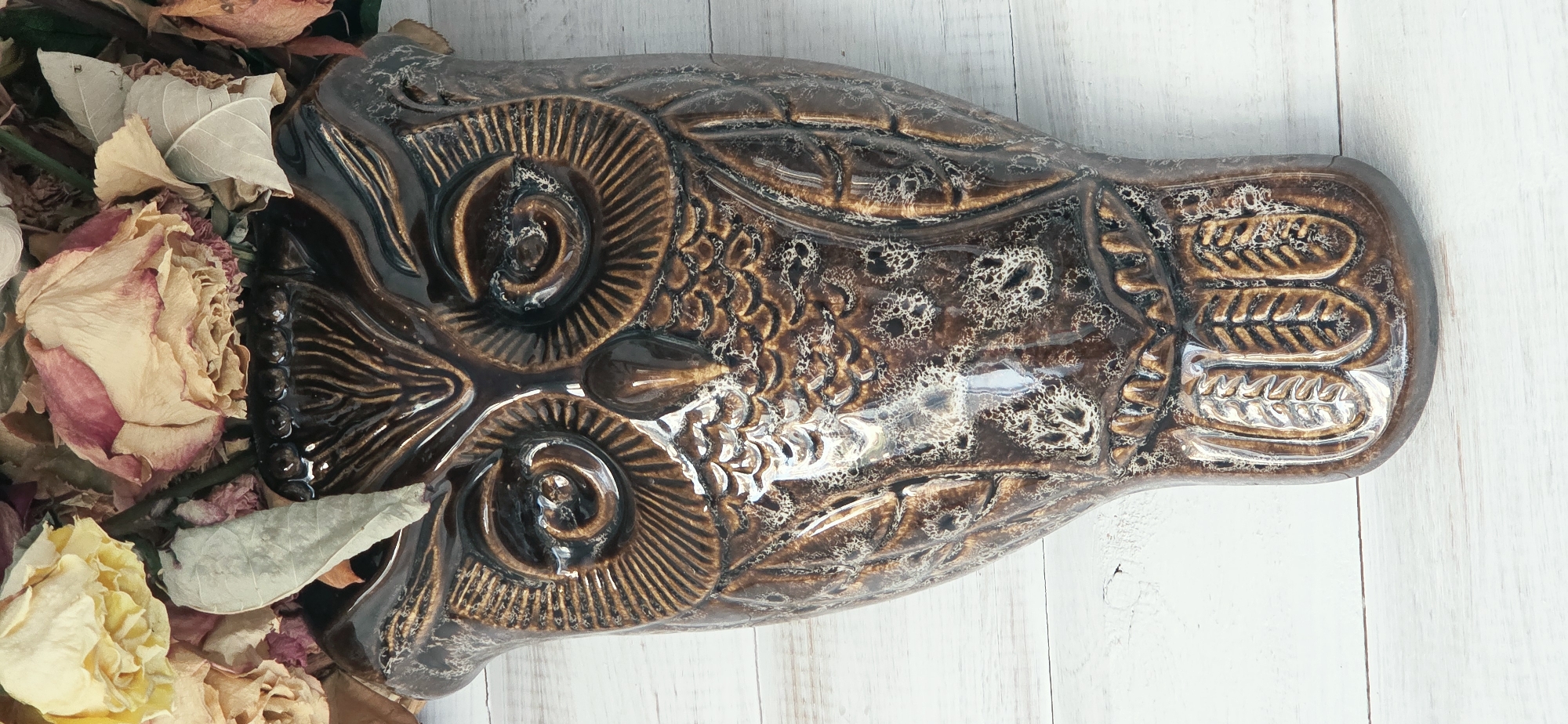 A close-up, eye-level shot shows a ceramic owl-shaped wall pocket filled with dried flowers. The owl is a dark brown with a glossy finish, featuring textured details for its eyes, beak, feathers, and facial markings. The dried flowers are a mix of yellow, pink, and beige roses, with some dried leaves interspersed. The owl is mounted against a background of white painted wooden planks. The image is well-lit, with the ceramic surface reflecting the light, creating highlights. There are no people or animals visible. The location in Lipetsk, Russia is not discernible from the image.