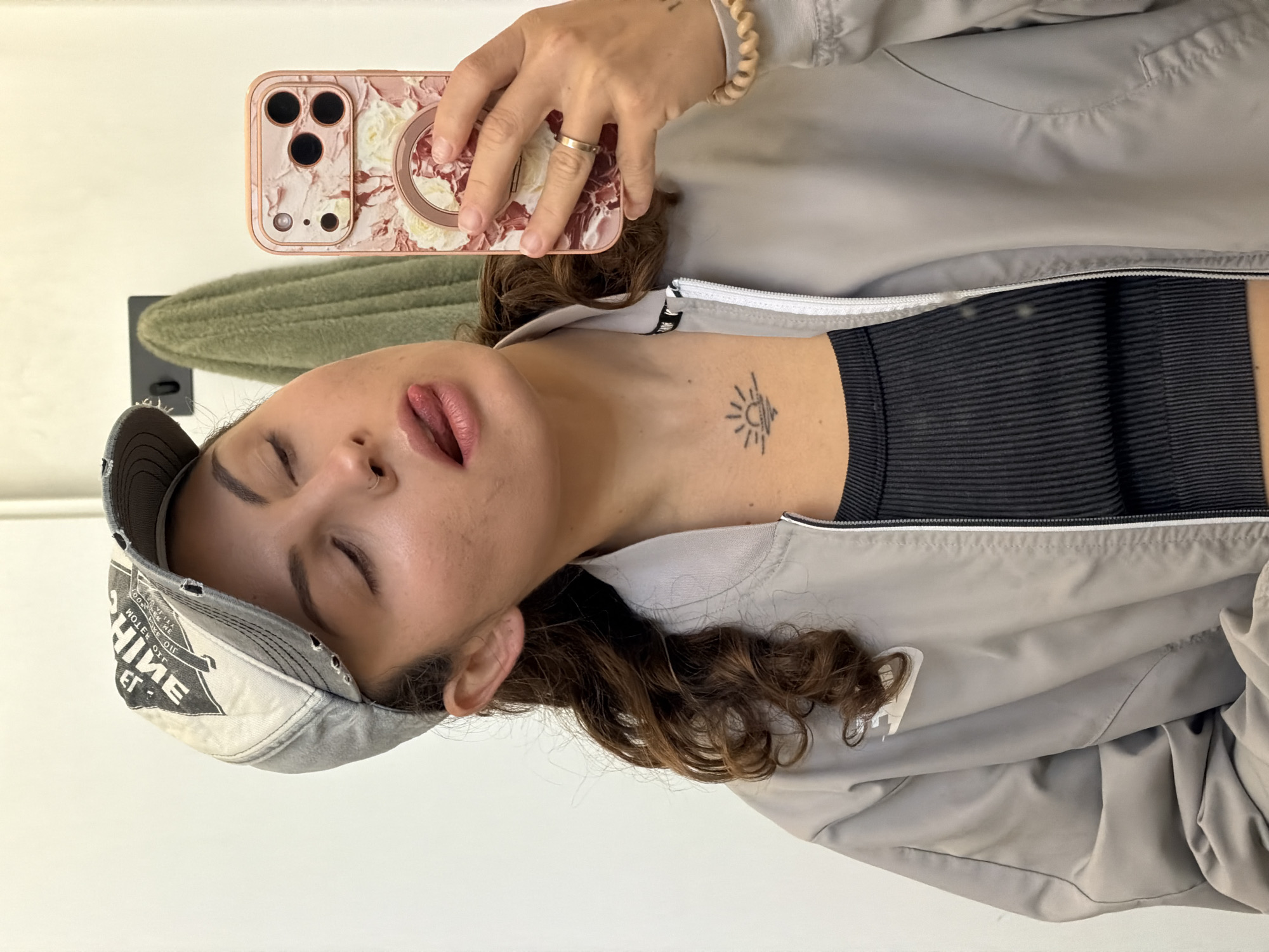 A woman is taking a selfie. She is wearing a gray baseball cap, a gray jacket, and a black top. Her hair is curly. She is holding a cell phone with a floral case. The woman has a tattoo on her neck of a sun. The location is unknown; there is no information in the image to suggest it is Kiryat Bialik, Israel.