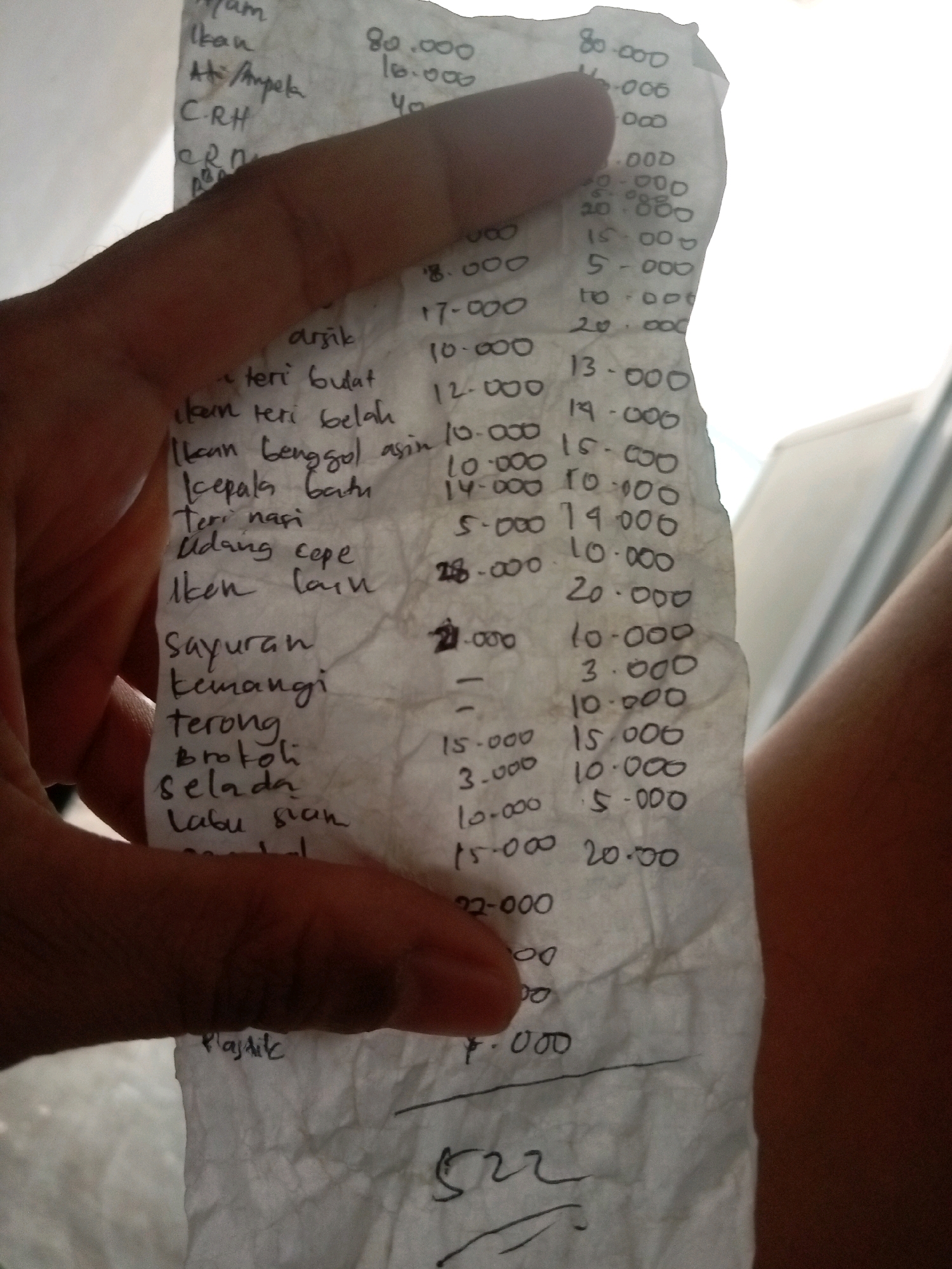 The image shows a close-up of a handwritten receipt on crumpled white paper, held by a person's hand in the foreground, partially obscuring the text. The receipt lists various food items and their prices. The writing is in Indonesian and appears to be from a local eatery or market. Based on the context of similar receipts and the commonality of handwritten sales slips in Indonesia, this scene is likely taking place in a market or food stall in Batam, Indonesia. The background is out of focus but suggests an indoor environment with bright lighting. Specific items listed include "Ikan" (fish), "Ayam" (chicken), "Sayuran" (vegetables), and "Udang cepe" (small dried shrimp), with corresponding prices such as 80.000, 16.000, and 5.000, likely in Indonesian Rupiah. There is also a line item for "Plastik" (plastic bag) at the bottom with a price of 1.000. The person's hand is brown-skinned and appears to be in motion, as indicated by the blur.