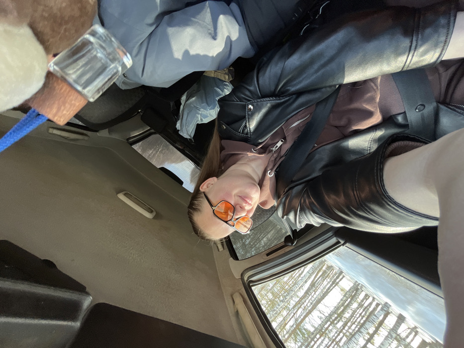 An image depicts the interior of a vehicle within the city of Nekrasovo, Russia. An adult female is positioned in the foreground, seated in what appears to be the front passenger seat. She has light brown hair, pulled back, and wears orange-tinted aviator-style sunglasses. Her expression is a slight smile directed towards the camera. She is dressed in a black leather-style jacket over a brown hooded sweatshirt and is secured by a black seatbelt across her chest and lap. Her left arm is extended towards the camera, suggesting she is taking a selfie.
To her right and slightly behind, the torso and legs of a second individual, wearing a grey or light blue jacket, are visible, likely occupying the driver's seat or another rear passenger seat. A light blue plastic bag is visible on a seat or floor in the mid-ground. Hanging from the overhead area of the vehicle, a clear glass bottle with a wooden top and a blue cord is partially visible. The vehicle's headliner is light grey and shows signs of wear.
Through the windows, a bright, sunny exterior environment is visible. This includes a dense collection of slender, light-colored tree trunks, indicative of a forest, possibly a birch grove, with what appears to be snow or frost on the ground, suggesting a winter or early spring season. The overall lighting is bright and natural.