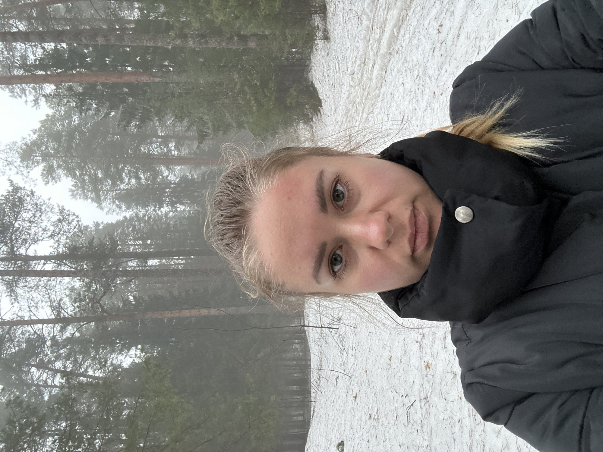 A selfie of a fair-skinned woman with blonde hair stands in a snowy area, likely a park or forest, with tall, bare trees visible in the misty background. She is wearing a puffy black jacket. The location is possibly Chaikovsky, Russia. The ground is covered in a layer of snow with some debris. The focus is on the woman's face, which is angled towards the camera.