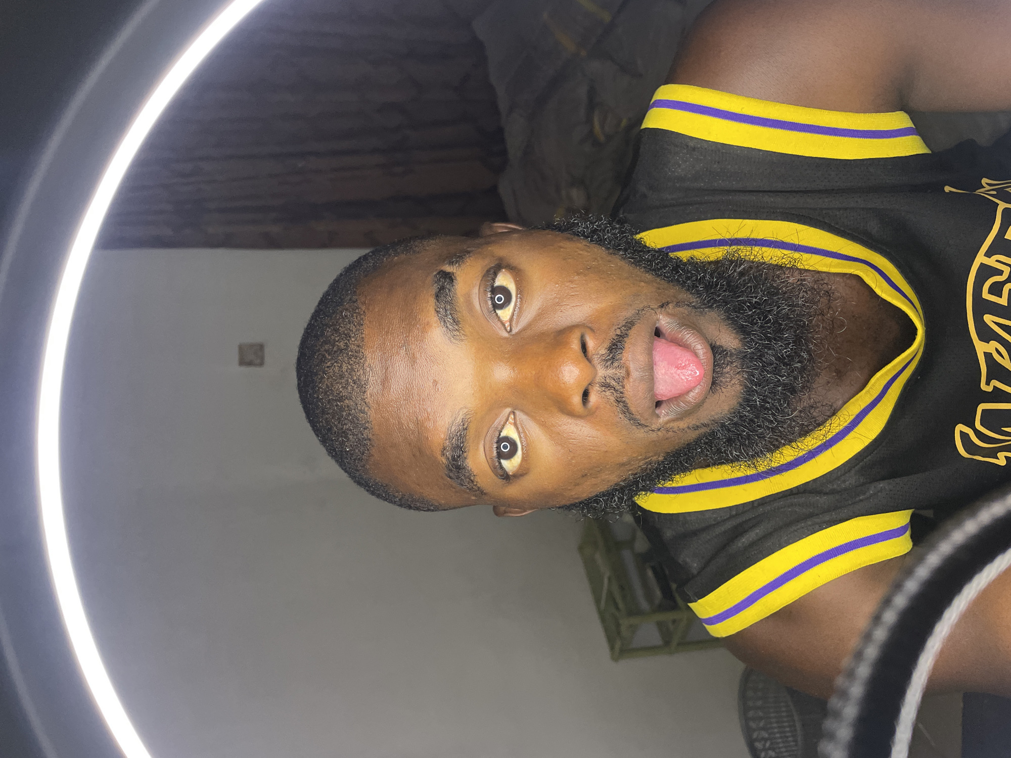 A close-up, medium shot shows a young Black man with a beard and short hair sticking his tongue out at the camera. He is wearing a black basketball jersey with yellow and purple trim. He is illuminated by a ring light, which creates a bright reflection in his pupils. The background is blurred and appears to be an indoor setting, possibly a bedroom, with a curtain and a small shelf visible. The scene is set in the city of Jos, Nigeria.