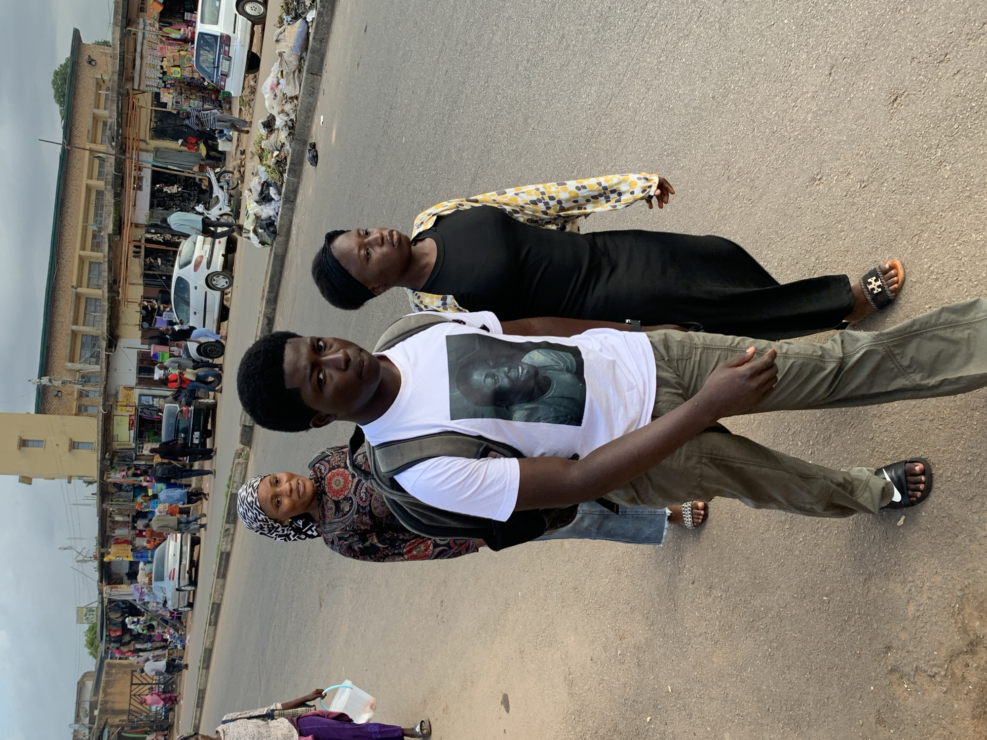 The image depicts a street scene in Jos, Nigeria, with a focus on three individuals walking on the right side of a paved road. The foreground features a young man wearing a white t-shirt with a portrait graphic and olive green pants, carrying a backpack. To his right is a young woman in a black dress and a patterned yellow and white over-shirt. Behind them, a woman with a headscarf and a patterned top is also walking.
The background reveals a bustling street lined with market stalls and shops on the left. Several cars, including a silver Peugeot and white vehicles, are visible on the road and parked along the curb. A motorcycle is also present. The street is moderately busy with other pedestrians in the distance. A pile of garbage and debris is situated along the curb to the right of the walking group. The sky is overcast, suggesting a cloudy day. The overall impression is that of an active urban marketplace environment.