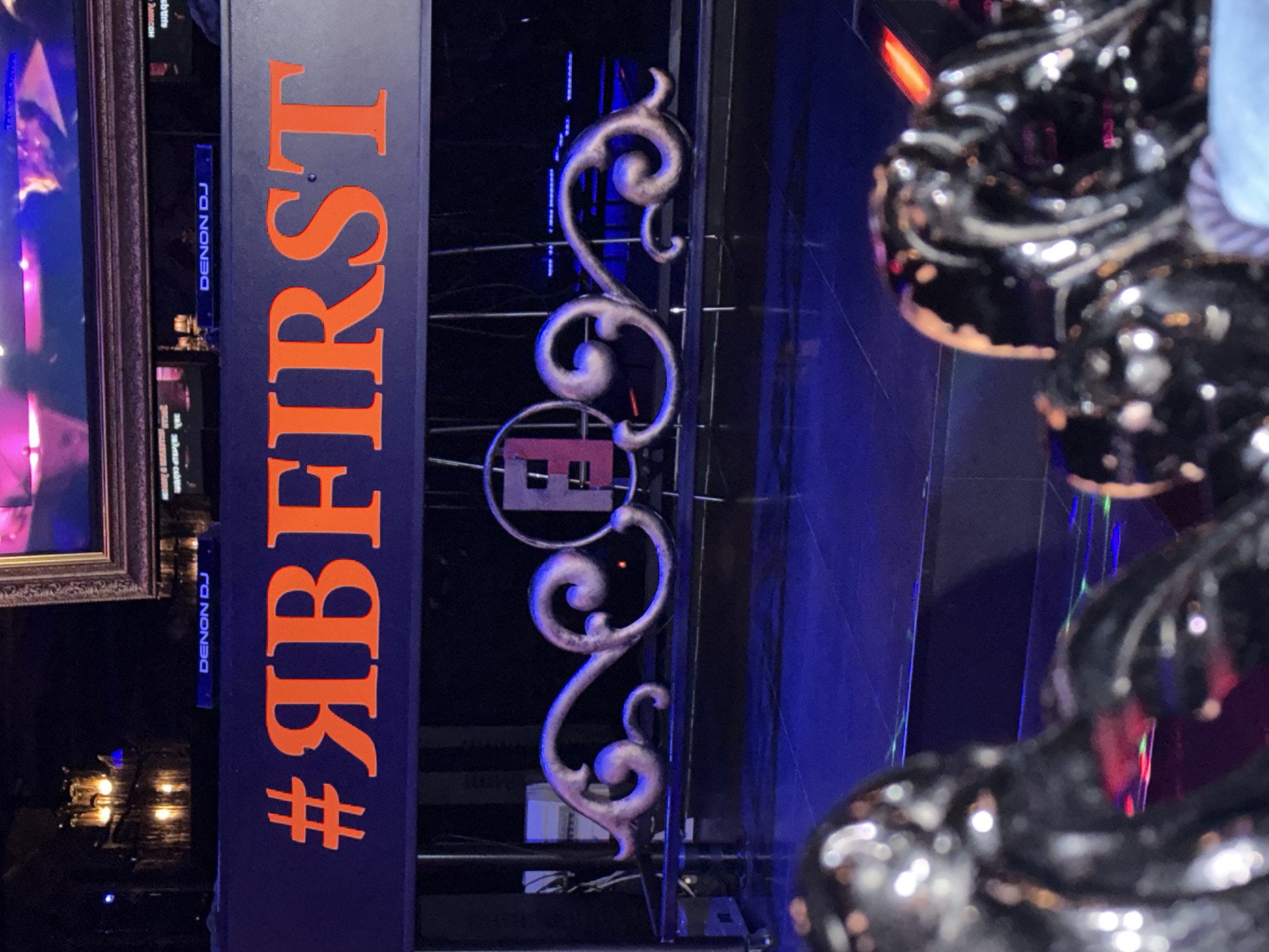 The image shows a vertical sign with "#ЯBFIRST" in orange lettering. "DENON DJ" is visible on the sign in a smaller font. A decorative wrought-iron design is positioned on the right of the sign. The background shows a blue-lit area, and a portion of a shiny, black decorative object is in the foreground on the right. No information is provided regarding the location, people, actions, interactions, or environment beyond what is visible in the image.