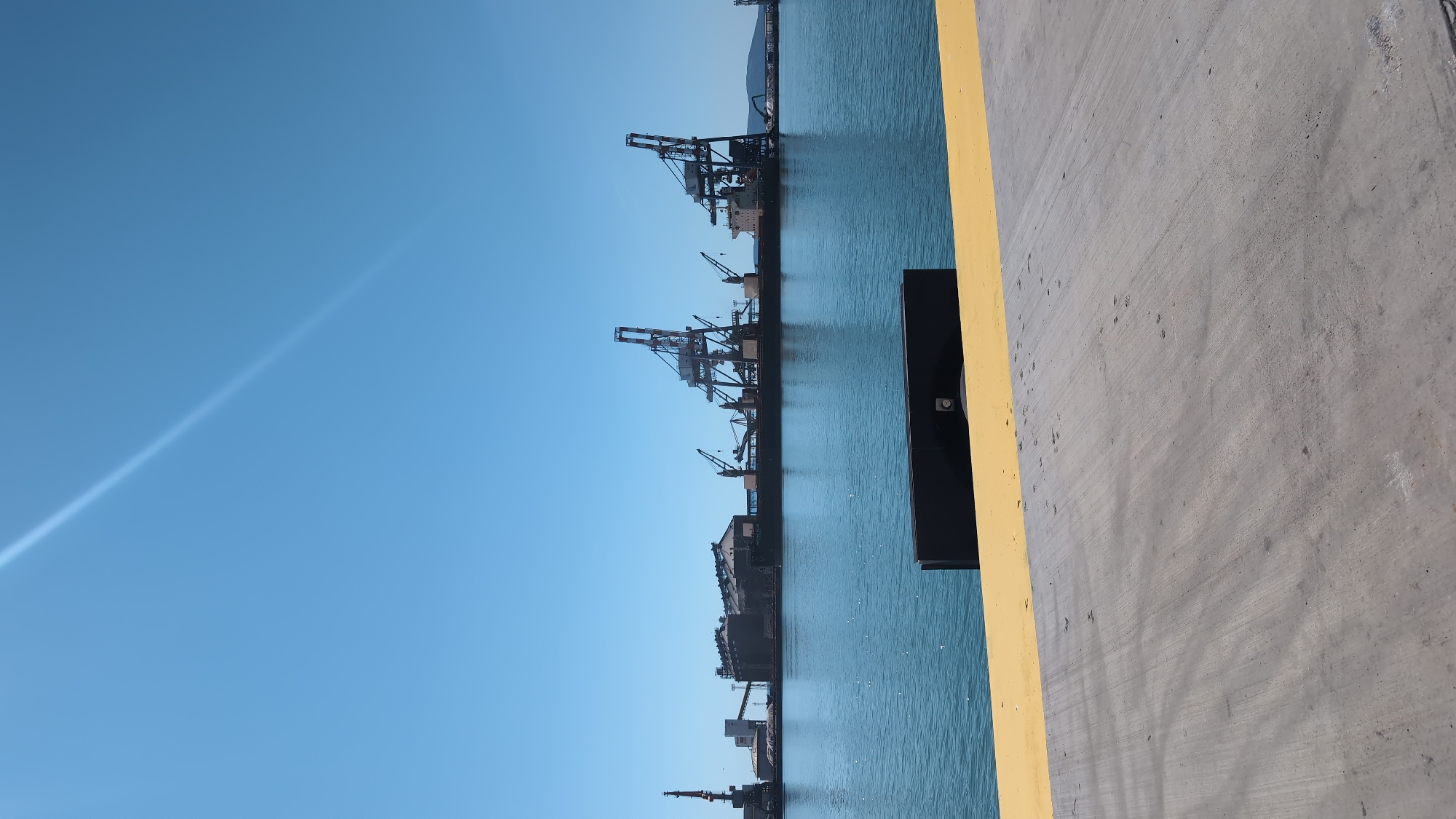 The image shows a wide view of a port area under a clear blue sky. In the foreground, a section of a concrete dock with a yellow painted edge is visible. The middle ground features a large body of blue water, likely a harbor or bay. Across the water, a long cargo ship is docked, laden with bulk materials. Numerous large cranes, characteristic of a port, are positioned along the ship and the shoreline. These include at least two prominent gantry cranes with red and white detailing, and several smaller cranes. Beyond the ship and cranes, industrial structures, possibly silos, are visible. In the distance, a hazy landmass or hills can be seen. The scene is bathed in bright sunlight, casting minimal shadows. The location is identified as the port of Burgas, Bulgaria, a major industrial and commercial hub on the Black Sea coast. There are no people or animals visible in the frame.