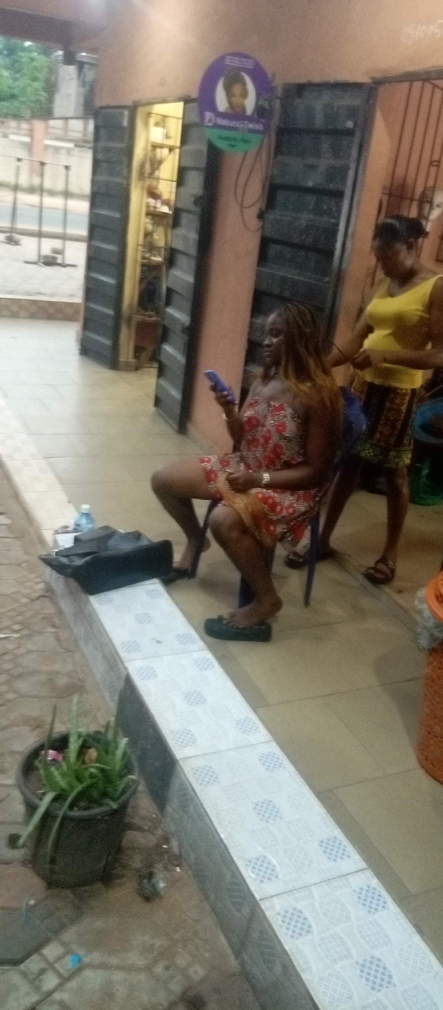 This image captures an outdoor hair styling session in Ogidi, Nigeria, likely in the late afternoon or early evening due to the soft, diffused lighting.
The main subjects are two women. One woman, with long brown dreadlocks or braids, is seated on a blue plastic chair, looking at a purple smartphone. She wears a red and white patterned strapless dress and black flip-flops. Her hair is being styled by a standing woman, who is positioned behind her. The standing woman wears a yellow sleeveless top and a patterned skirt, and her hair is tied back.
They are situated outside an open shop entrance. The shop doorway has metal slatted doors pushed open, revealing shelves with various products inside. Above the doorway hangs a circular sign, predominantly purple and green. The text visible on the sign reads "DARLING" at the top, with an illustration of a woman's head with curly hair, and "Natural Twish" below it.
In front of the seated woman, a tiled ledge or barrier is present, on which a black bag and a clear plastic water bottle are placed. The ground is paved with interlocking concrete blocks in the foreground, transitioning to a smoother tiled surface near the women. A black potted plant with green foliage sits on the ground to the left. The overall setting appears to be a commercial area or roadside business. The seated woman appears relaxed, engrossed in her phone while her hair is being worked on.