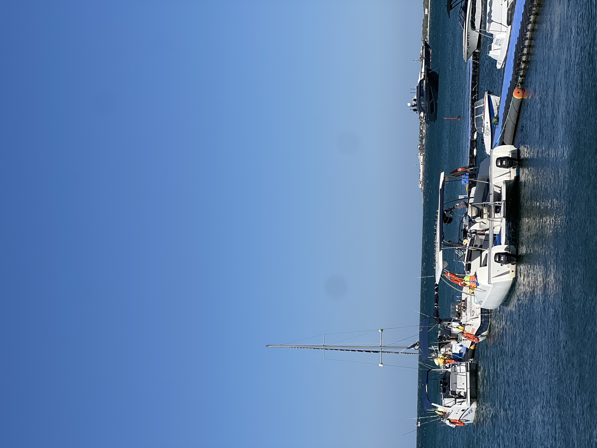 A bright, clear day with a deep blue sky over a calm, teal-colored sea. In the foreground, a series of docked boats are tied to a blue floating dock. The boats are of various sizes and types, including several small white motorboats, a white catamaran with a blue canopy, and a white sailboat with a tall mast. Some of the motorboats have fishing rods extending from them. In the middle ground, a sleek, dark-colored yacht is anchored in the water. In the background, a distant coastline is visible with buildings and structures. The scene appears to be a marina or harbor.