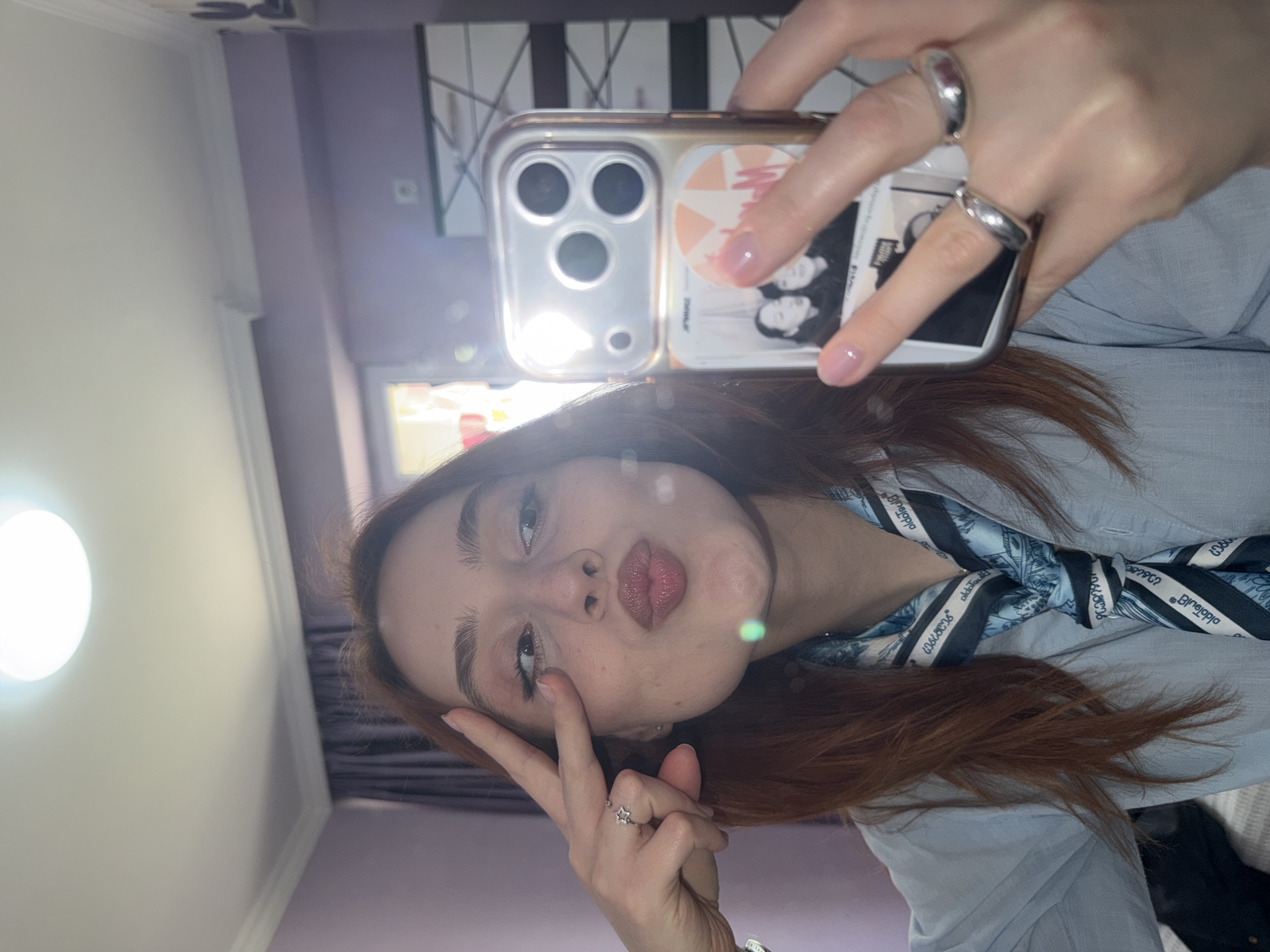 A woman is taking a selfie in an indoor location with what appears to be a phone. She is wearing a light blue shirt with a patterned scarf tied around the neck. The woman has brown-red hair and is wearing several rings on her fingers. The phone has a case with a design on it. The location cannot be determined from the provided image.