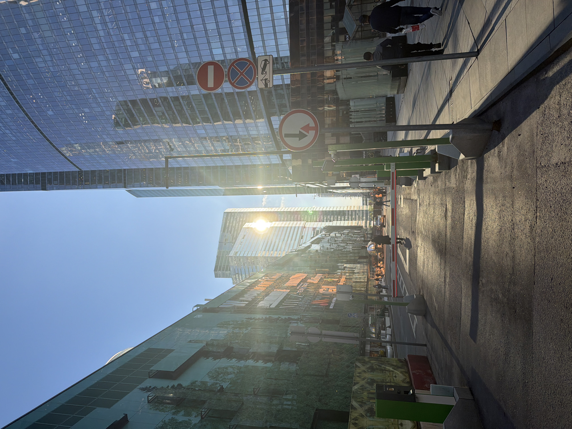 The image depicts a street scene in Khimki, Russia. Tall, modern glass buildings line the street, reflecting sunlight. Several street signs are visible, including a "no entry" sign and a "one-way street" sign. Pedestrians are walking along the sidewalk. The street is paved and appears to be wet. The sky is clear and blue.