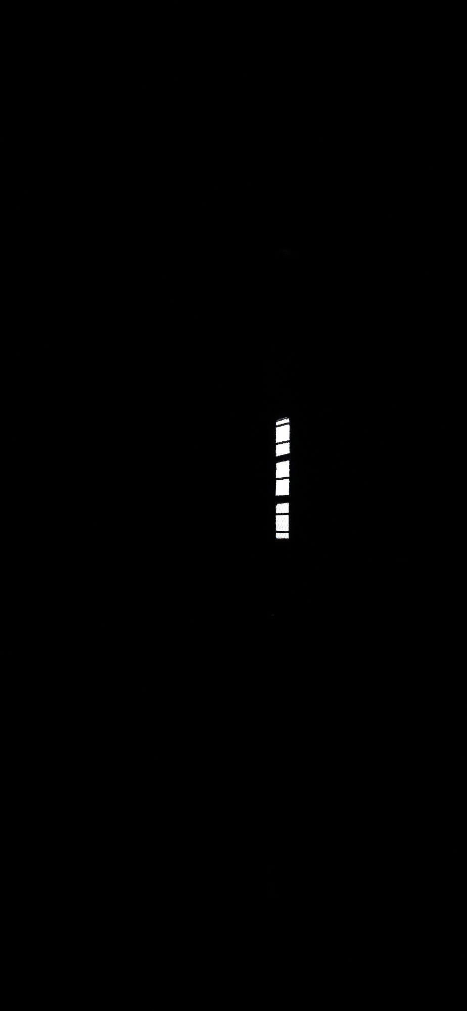 The image is a black and white photograph that appears to be taken inside a building. The majority of the image is black, indicating a dark environment. On the right side of the image, there is a vertical arrangement of several white rectangular shapes. These shapes are thin and evenly spaced, resembling the panes of a window or ventilation slats. There is no visible indication of the location within Port Blair, India, or any people, subjects, actions, or interactions. The environment appears to be a dimly lit interior space.