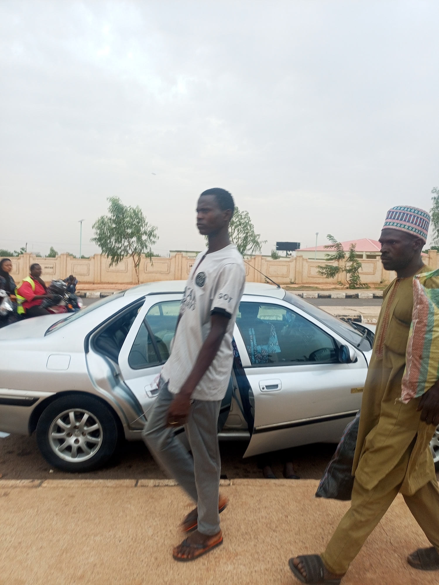 The image captures an urban scene in an unspecified city. Two men are walking along a sidewalk, with a silver sedan parked nearby, its rear passenger door open. One man wears a light-colored t-shirt and grey pants, while the other wears traditional yellow clothing and a patterned hat. In the background, two individuals on a motorcycle wait on the roadside. A low wall and some buildings can be seen beyond the road. The sky is overcast, suggesting muted daylight conditions.